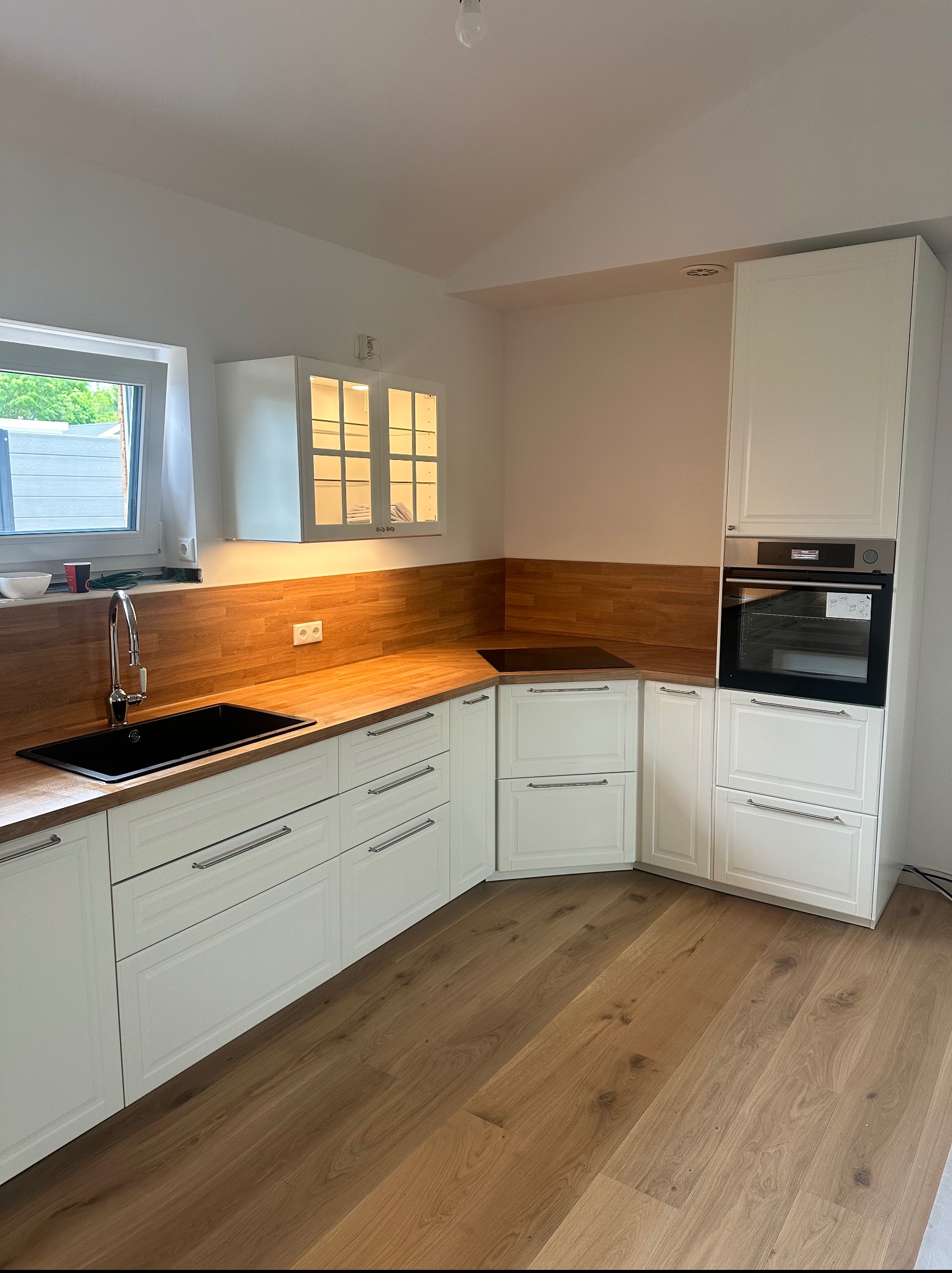 Nowoczesna kuchnia z białymi szafkami, drewnianym blatem i podłogą. Widoczny zlew, płyta indukcyjna i piekarnik w zabudowie. Górna szafka z podświetlanymi szybkami.