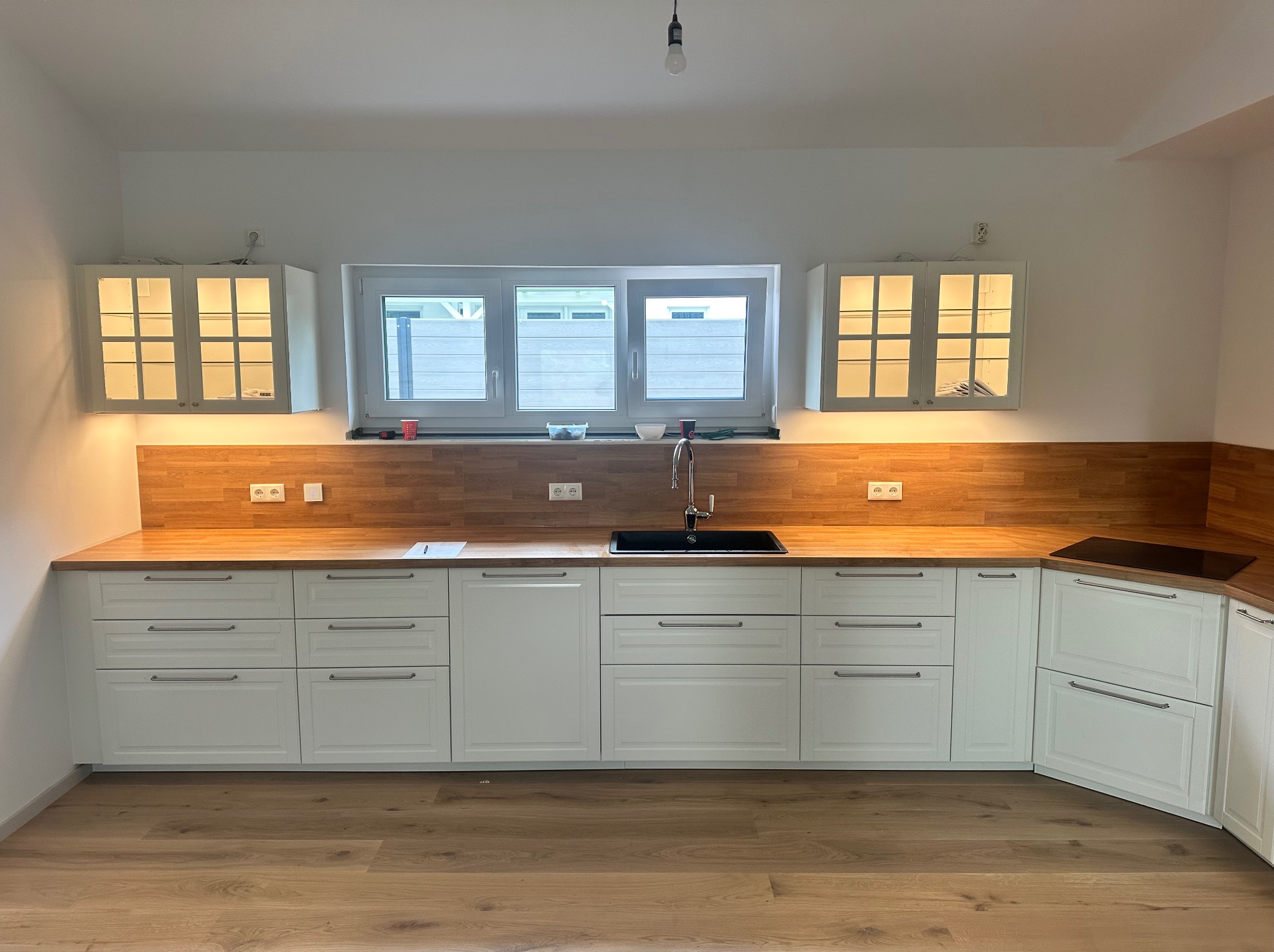 Jasna kuchnia z drewnianym blatem i oświetlonymi szafkami wiszącymi. Minimalistyczny design, duży zlew i płyta indukcyjna. Nowoczesna aranżacja wnętrza.