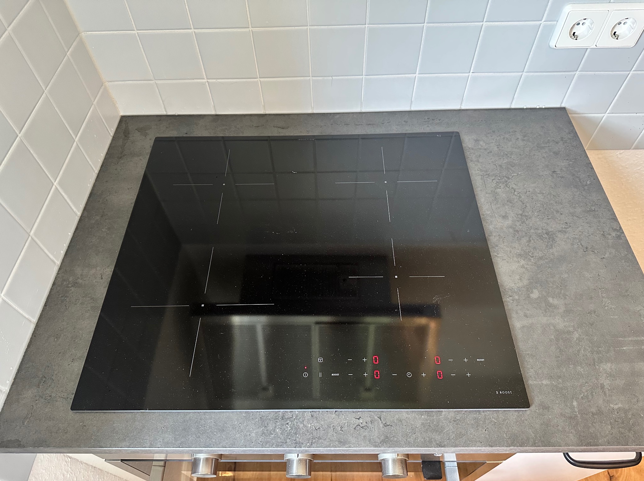 Czarna płyta indukcyjna marki Brolly wkomponowana w szary blat kuchenny, z białymi, kwadratowymi płytkami w tle, widok z góry.