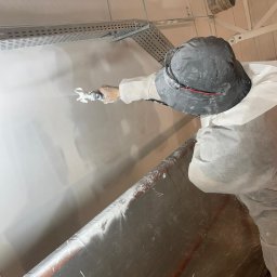 Mateusz Kupiec - Pracownik w kombinezonie ochronnym nanosi gładź natryskową na ścianę w pomieszczeniu z metalową konstrukcją sufitową. Widoczny pistolet natryskowy i wiadro z gładzią.