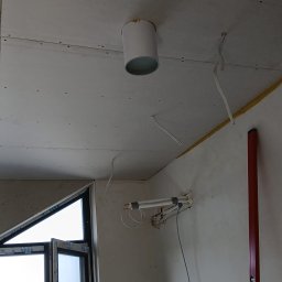 Mateusz Kupiec - Montaż płyty gipsowo-kartonowej na suficie z widocznymi przewodami i elementami instalacji. W tle okno z widokiem na zieleń. Czerwona poziomica oparta o ścianę.