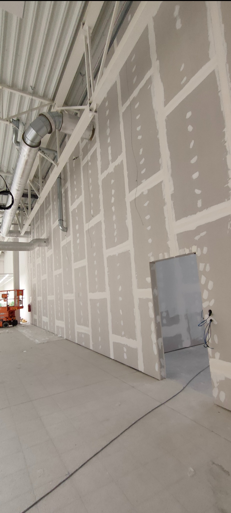 Wnętrze w trakcie prac wykończeniowych: ściany z płyt gipsowo-kartonowych, widoczne łączenia szpachlowane, sufit z instalacją wentylacyjną. W tle podnośnik.