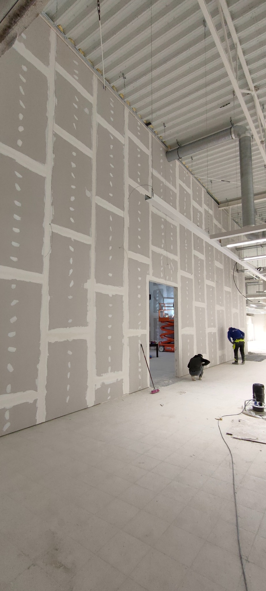 Montaż płyt gipsowo-kartonowych na ścianie w dużym pomieszczeniu. Widoczni dwaj pracownicy podczas pracy. W tle podnośnik. Oświetlenie sufitowe.