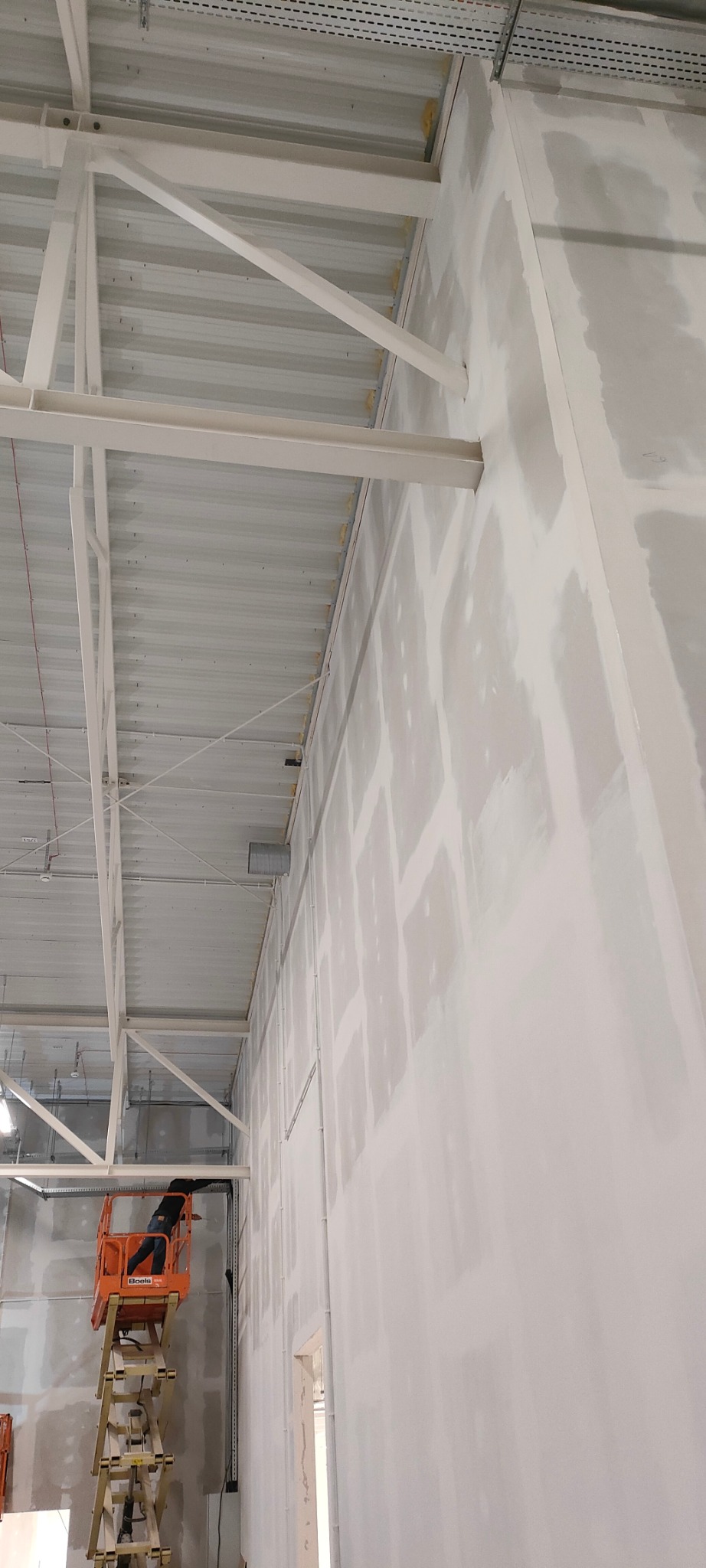 Praca na wysokości: pracownik na podnośniku maluje ścianę wewnątrz hali. Widoczne metalowe elementy konstrukcyjne i sufit. Ściana przygotowana do dalszych prac wykończeniowych.