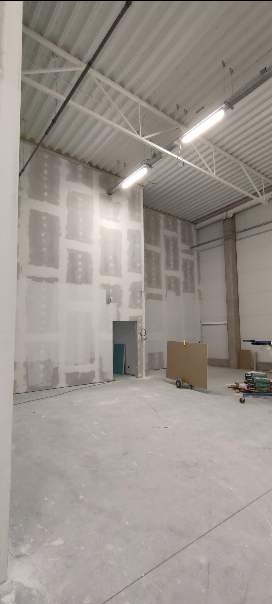 Montaż płyt karton gips w hali przemysłowej. Widoczne łączenia płyt przygotowane do szpachlowania. Na podłodze leżą narzędzia i materiały budowlane.