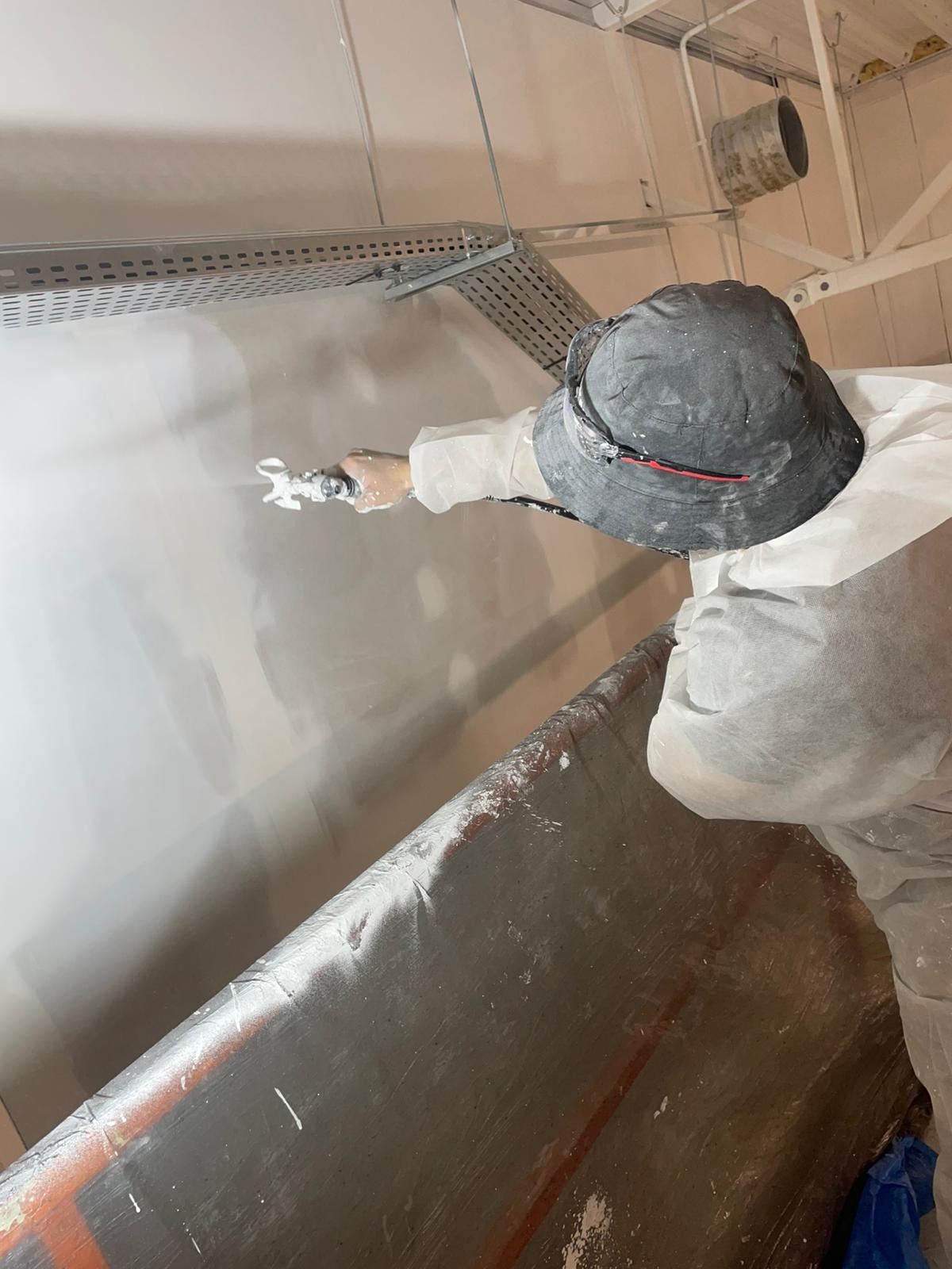 Pracownik w kombinezonie ochronnym nanosi gładź natryskową na ścianę w pomieszczeniu z metalową konstrukcją sufitową. Widoczny pistolet natryskowy i wiadro z gładzią.