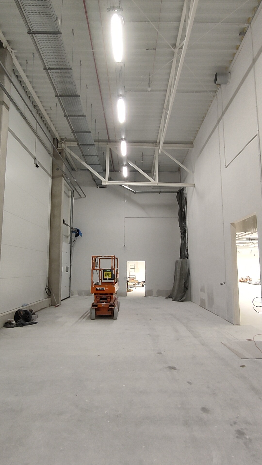 Wnętrze hali w trakcie prac wykończeniowych. Widoczny podnośnik koszowy, białe ściany, sufit z instalacjami i oświetleniem. W tle otwarte przejście do kolejnej przestrzeni.