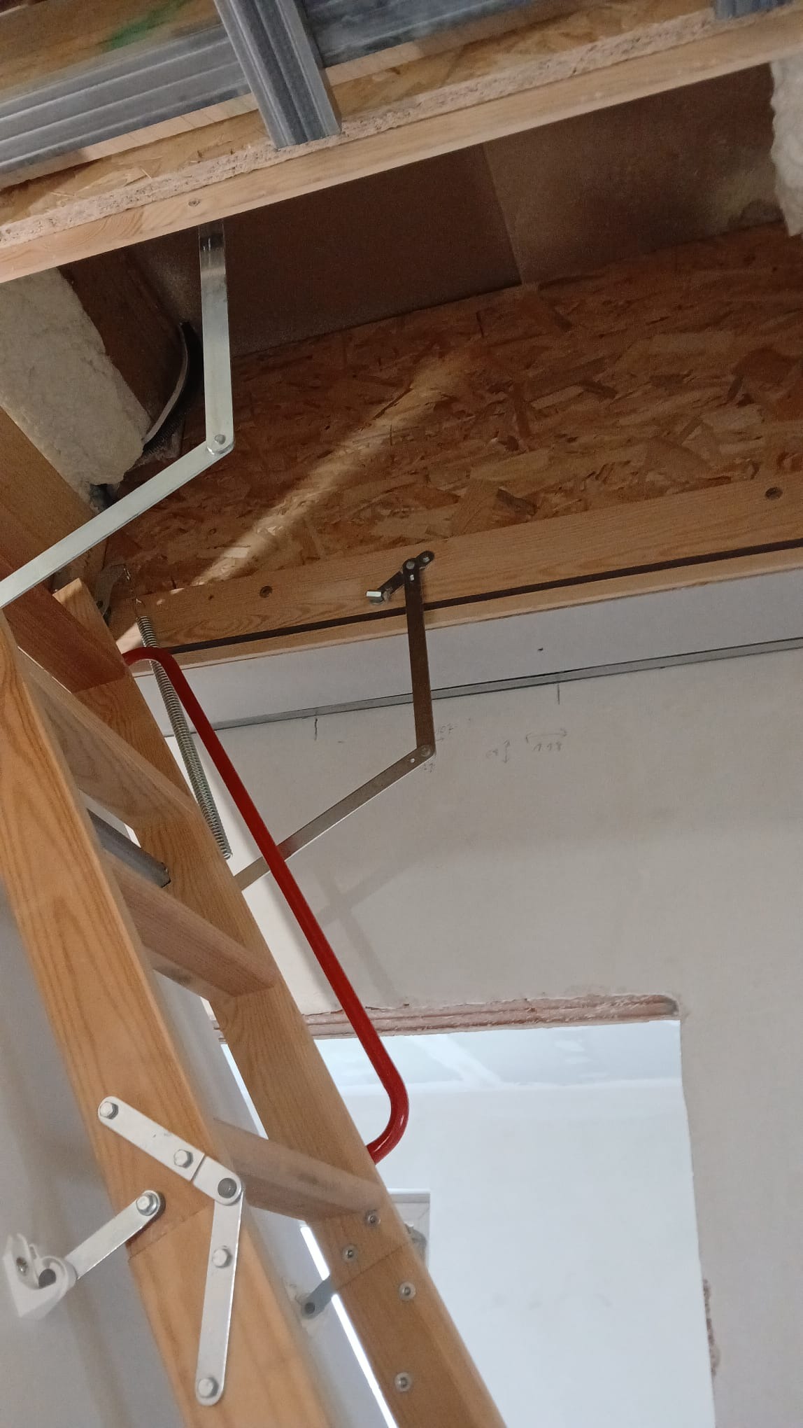 Drewniane schody strychowe z czerwonym uchwytem, zamontowane w suficie z widoczną konstrukcją metalową i płytą OSB. Widoczny fragment ściany z otworem.