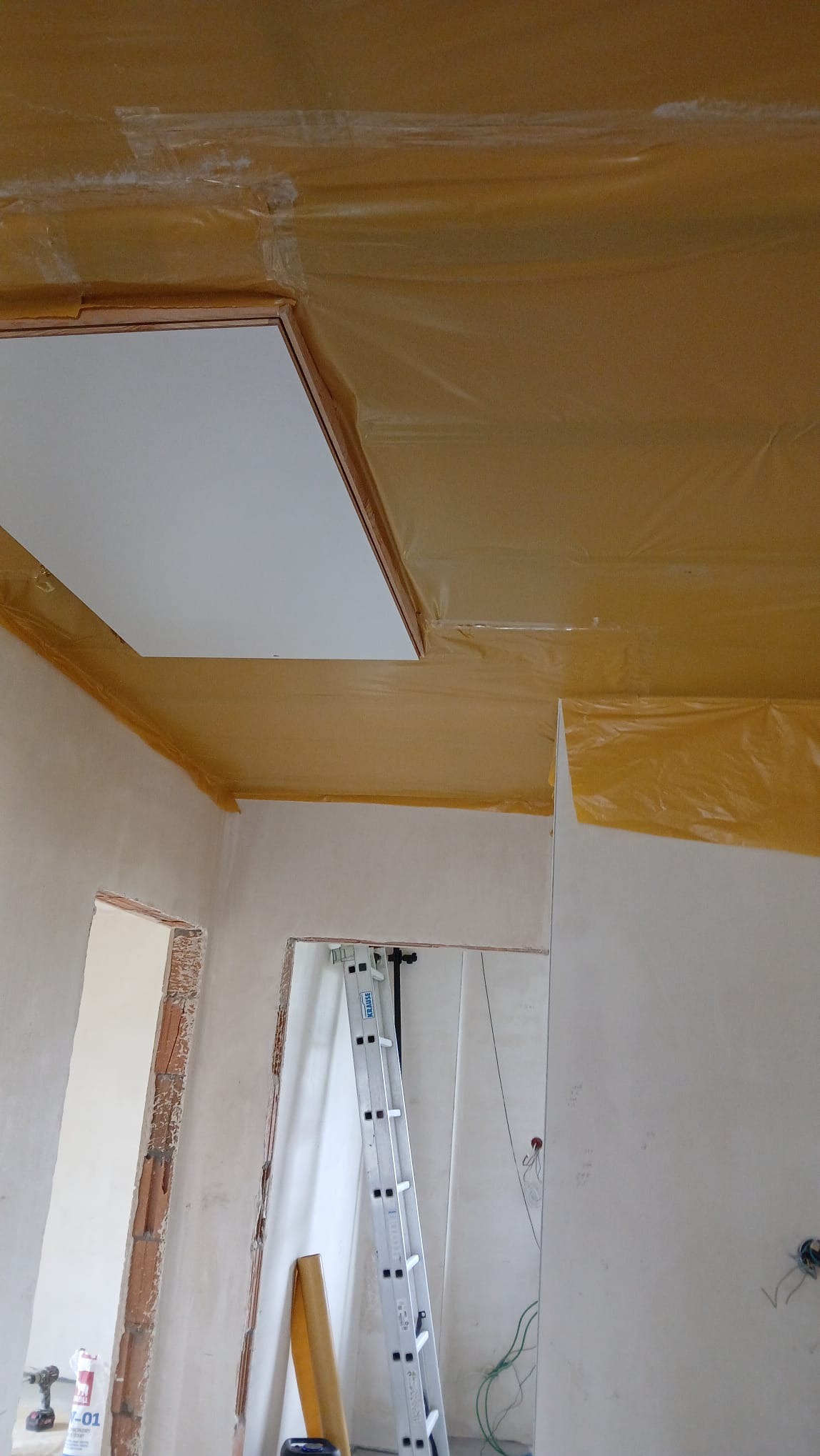 Przygotowane pomieszczenie do prac wykończeniowych: sufit i ściany zabezpieczone folią, widoczna drabina i otwory drzwiowe w stanie surowym.