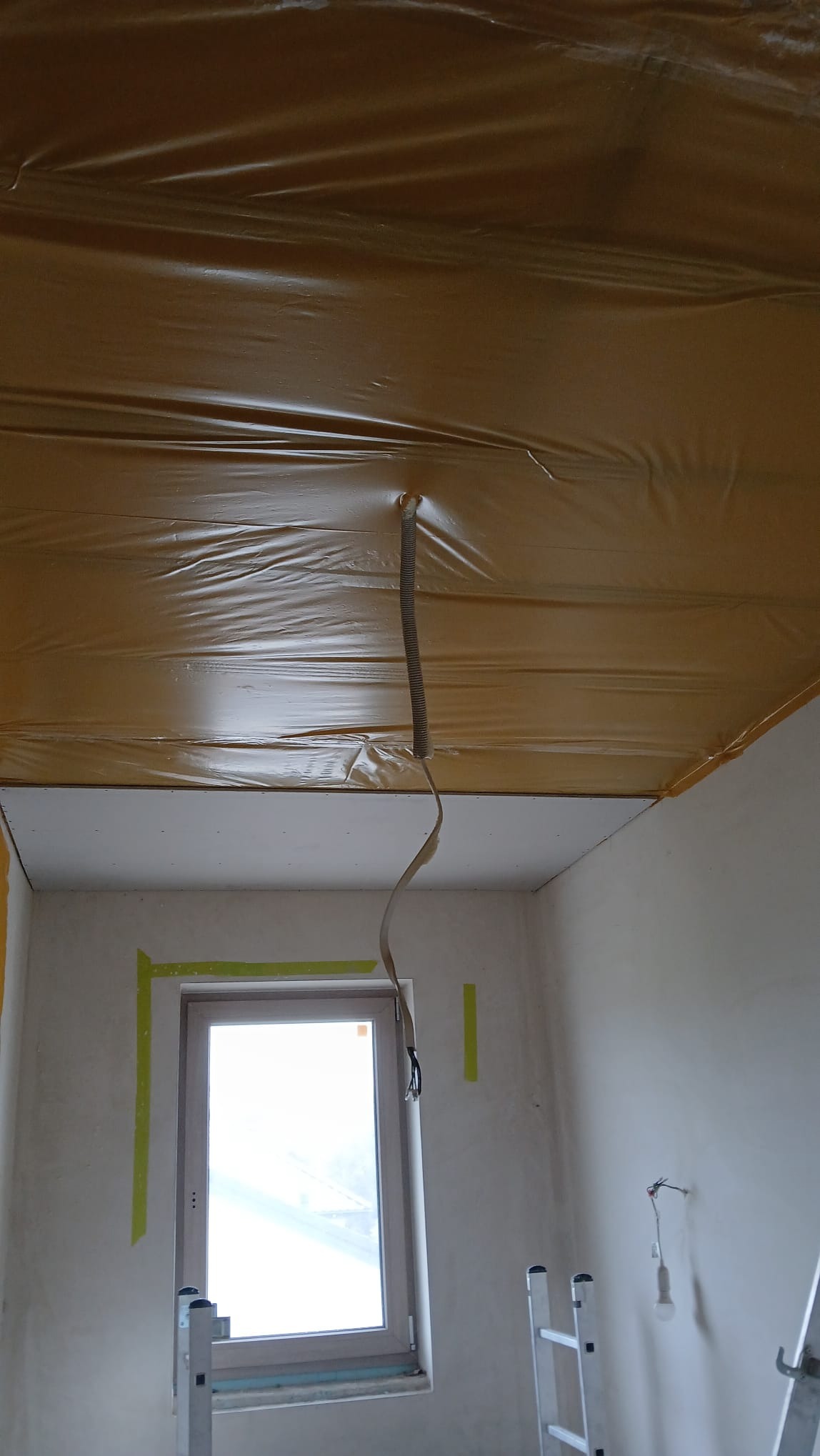 Przygotowanie do montażu sufitu podwieszanego: folia ochronna na suficie, widoczne profile i taśmy zabezpieczające okno. Etap prac wykończeniowych.