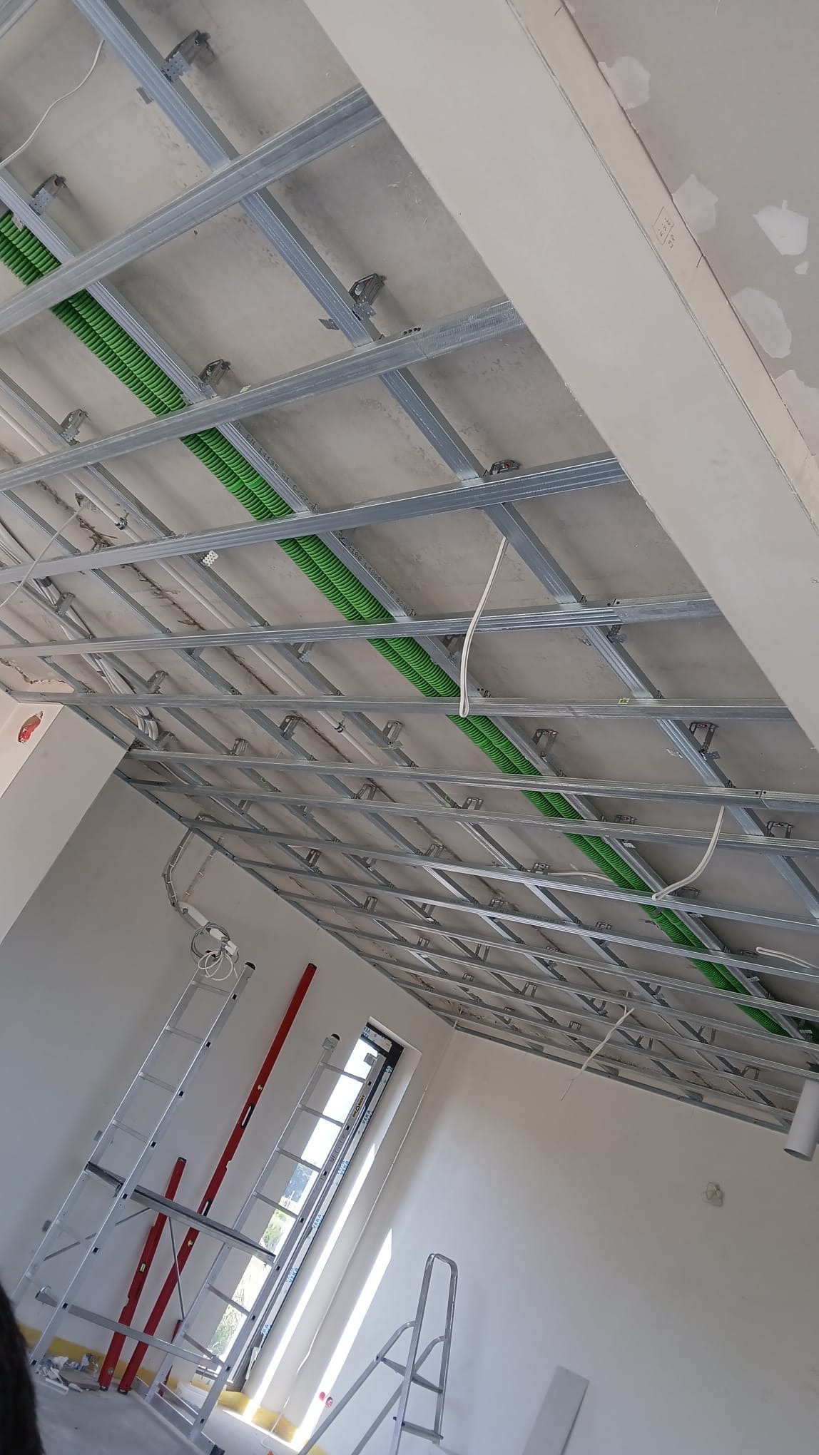 Sufit podwieszany z metalową konstrukcją i zielonymi rurami instalacyjnymi, widok z dołu, drabina i poziomica w tle, surowy stan wykończenia.