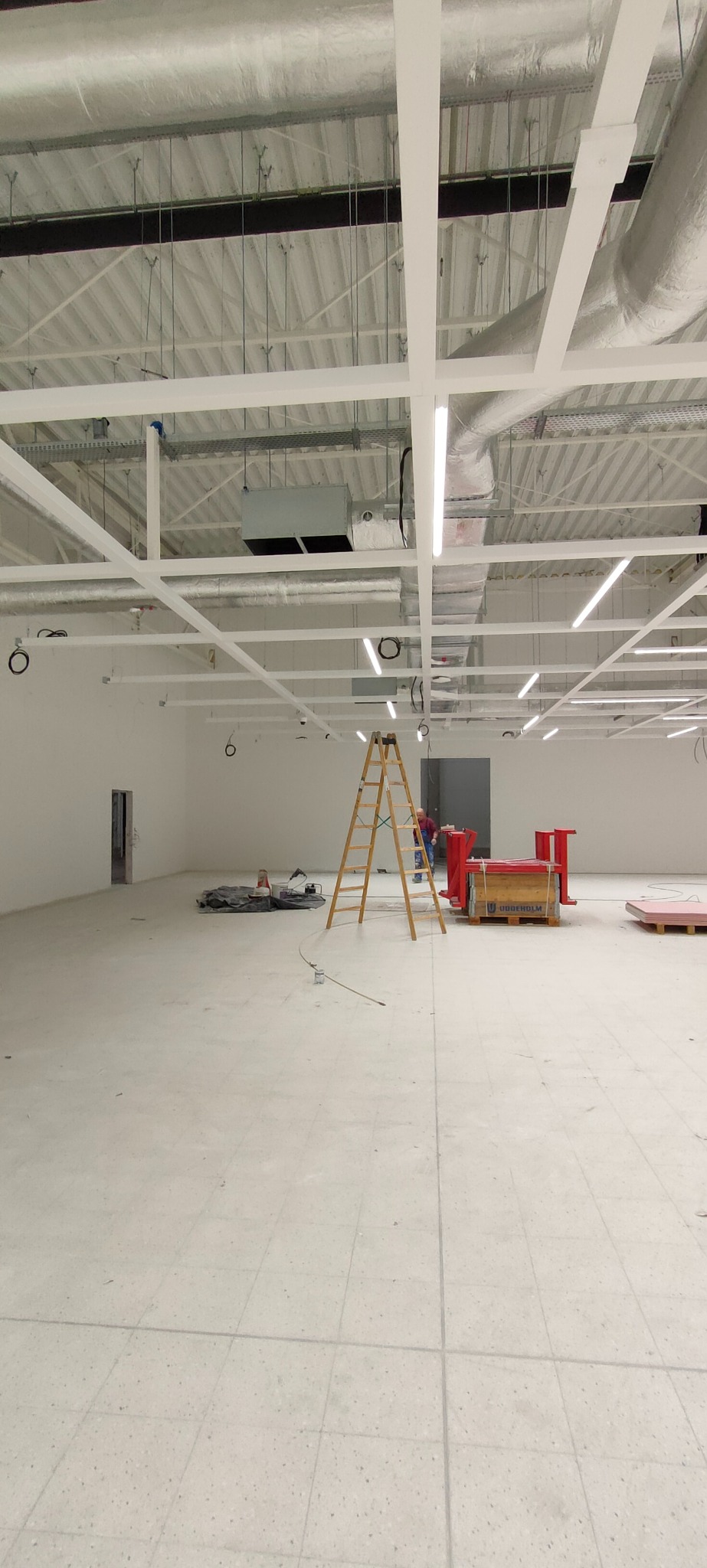 Wnętrze w trakcie wykończenia: surowy sufit z instalacjami, drabina, materiały budowlane. Widoczny fragment osoby w oddali. Duża, pusta przestrzeń przygotowana do prac.