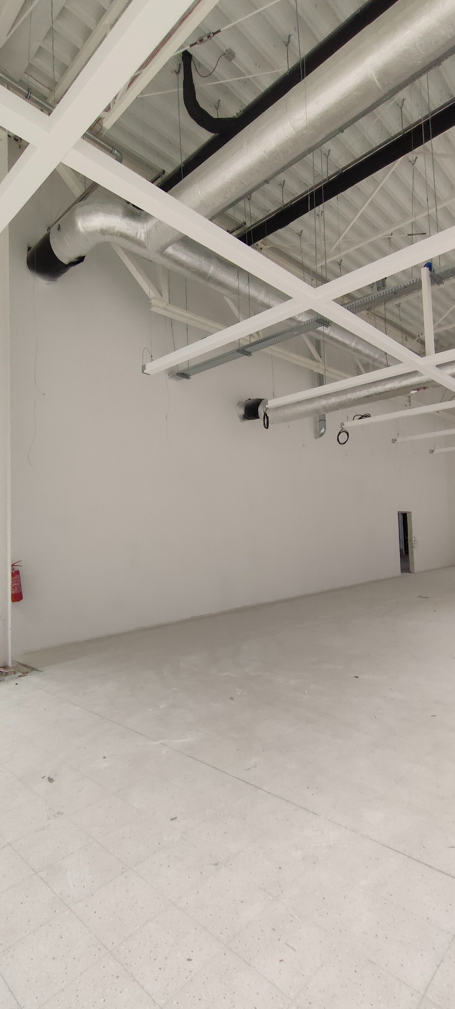 Wnętrze hali w trakcie wykańczania: białe ściany, sufit z instalacjami, posadzka z płytek. Widoczny gaśnica i otwór drzwiowy. Perspektywa z niskiego kąta.