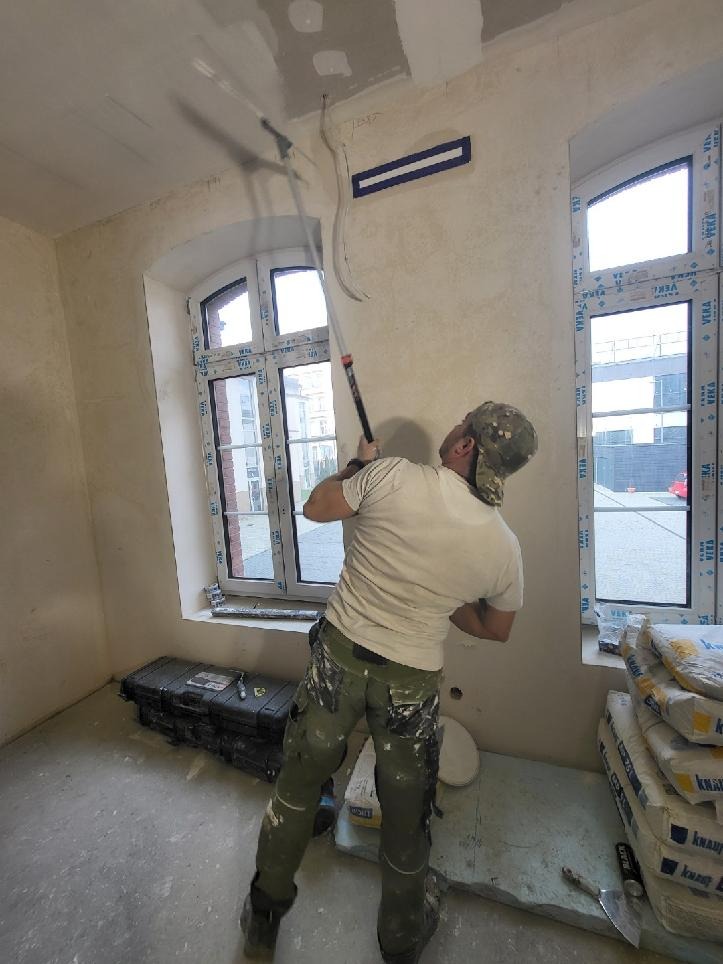 Mężczyzna maluje sufit wałkiem teleskopowym w jasnym wnętrzu z oknami. Widoczne worki z zaprawą i narzędzia malarskie. Praca w toku.