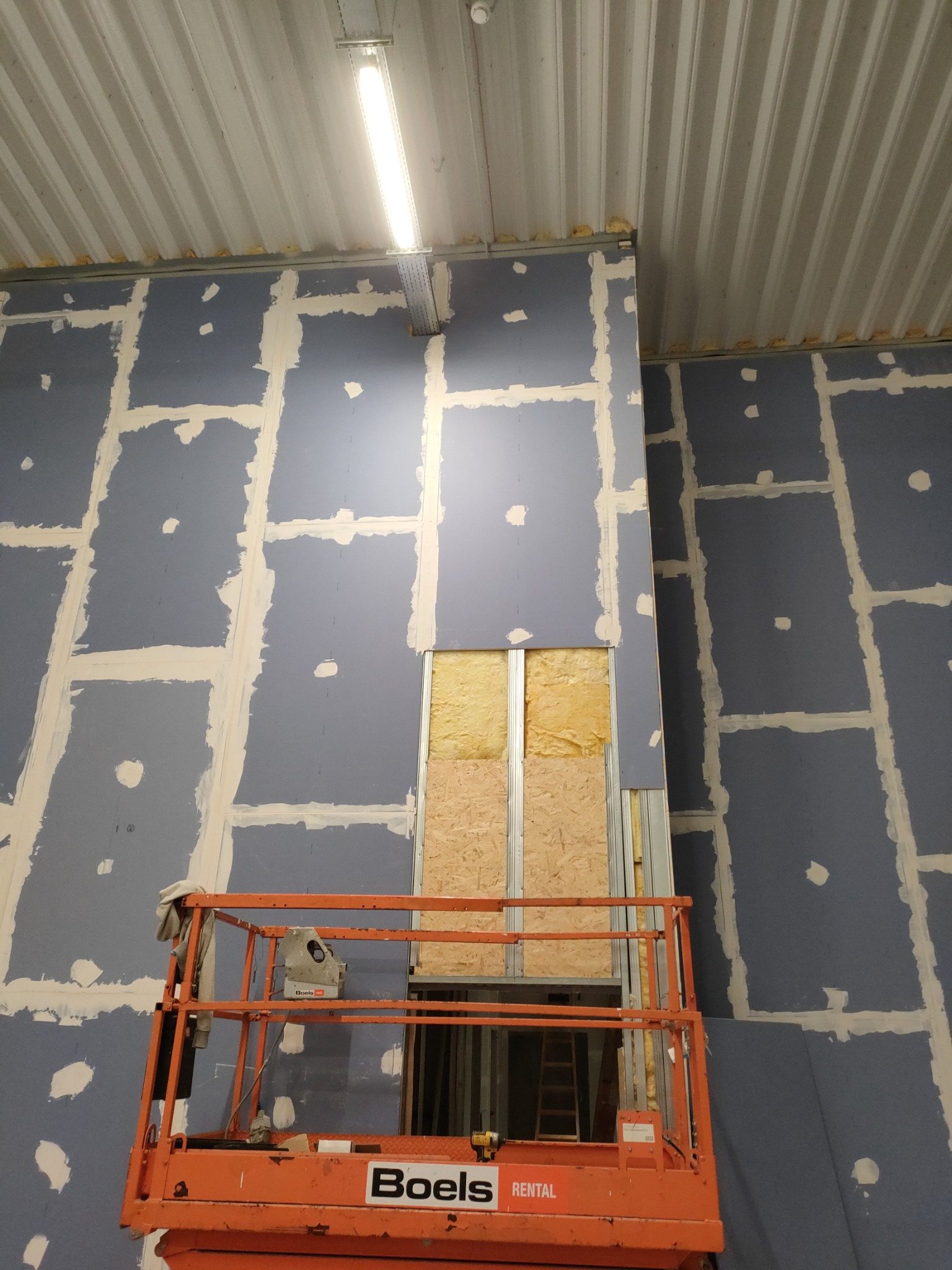Montaż płyt gipsowo-kartonowych na ścianie w hali, widoczna konstrukcja z izolacją, podnośnik Boels Rental. Etap szpachlowania łączeń płyt.