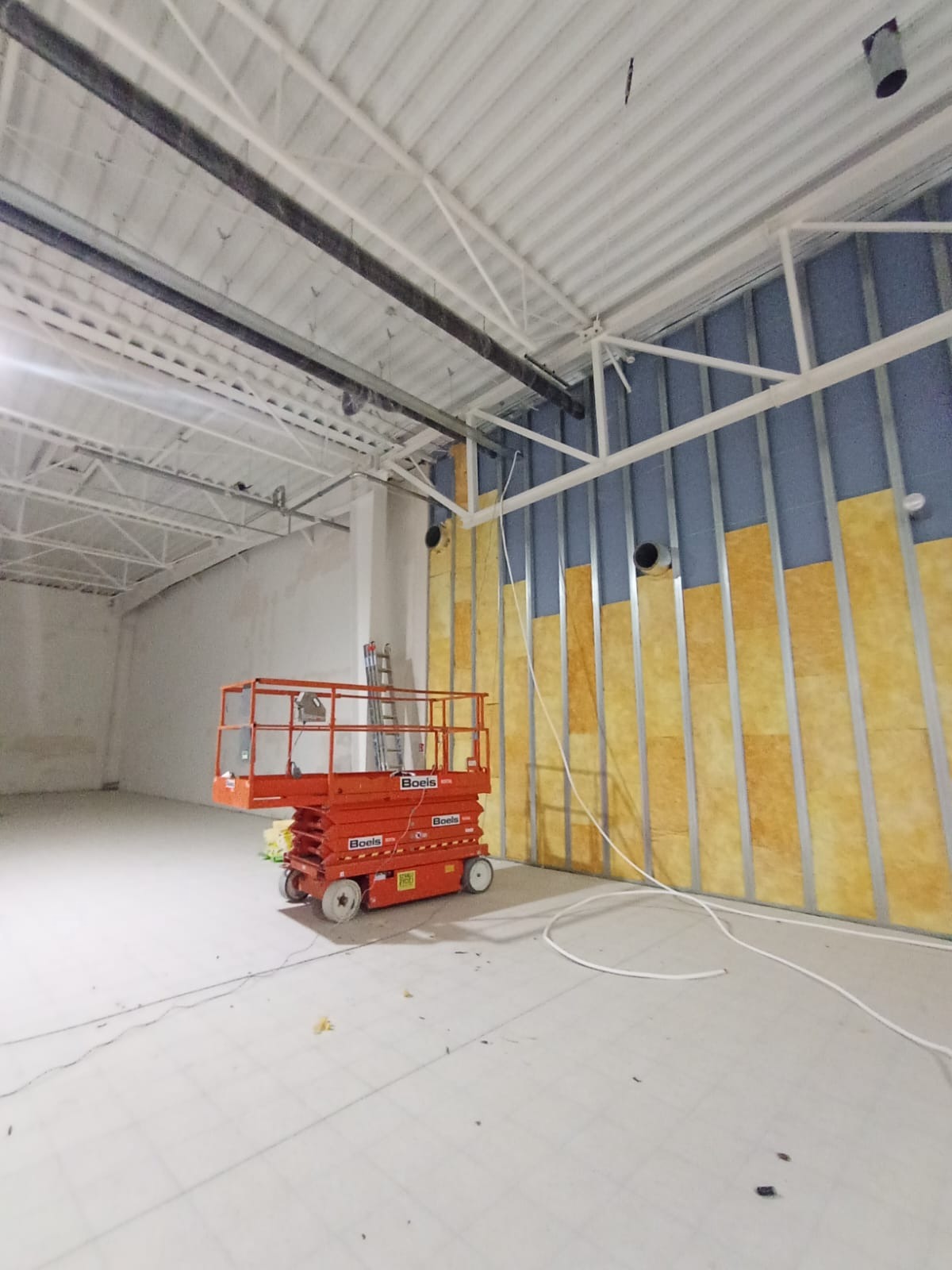 Montaż izolacji w budynku komercyjnym: stalowa konstrukcja, wełna mineralna, podnośnik Boels. Widoczne rury pod sufitem i przewody na podłodze.
