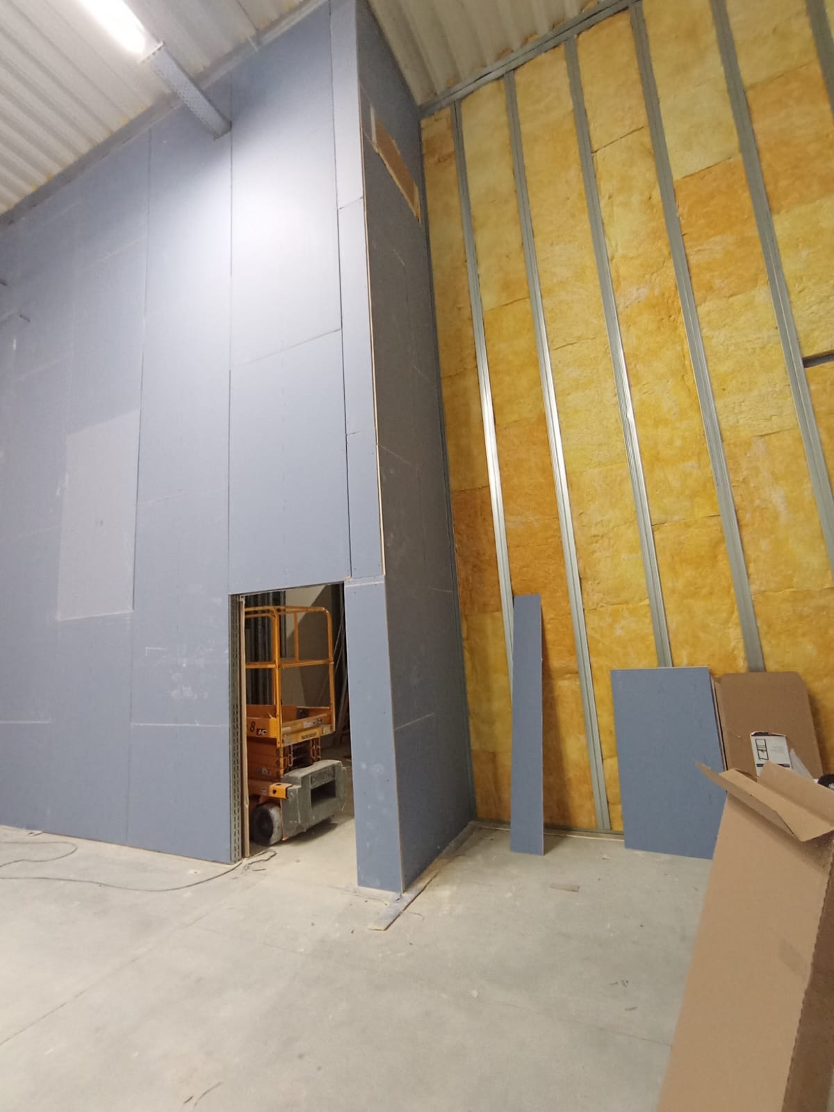 Wnętrze w trakcie prac wykończeniowych: ściana z płyt karton-gips, widoczna izolacja z wełny mineralnej, podnośnik. Częściowo zakryte kartony na podłodze.