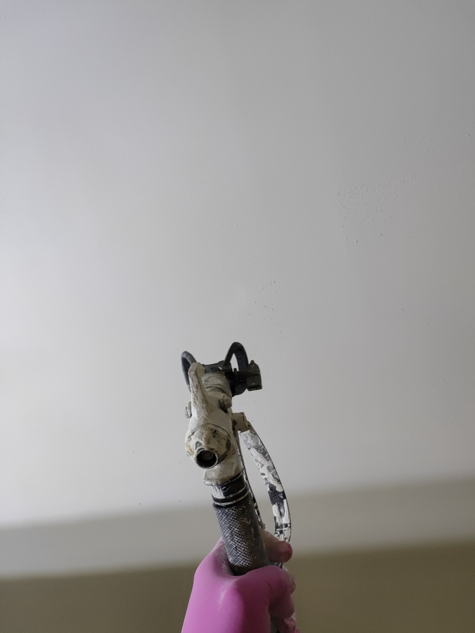 Malowanie natryskowe sufitu z bliska. Pistolet w dłoni w różowej rękawicy, widoczne drobne kropelki farby na świeżo pomalowanej powierzchni.