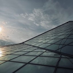 Kamil Chadaj Frat - prace wysokościowe - Pracownik na szklanym dachu budynku, zabezpieczony linami. Widok z dołu, słońce odbija się w szybach, pochmurne niebo w tle. Praca na wysokości.