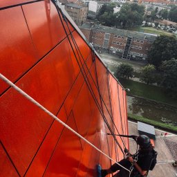 Kamil Chadaj Frat - prace wysokościowe - Pracownik w kasku, na linach, czyści czerwoną elewację budynku z użyciem myjki ciśnieniowej. W tle panorama miasta z rzeką i zabytkowymi budowlami.