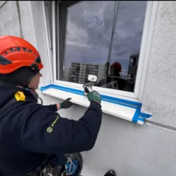 Pracownik w kasku i uprzęży myje okno na wysokości, zabezpieczone taśmą. Na szybie odbija się budynek i pochmurne niebo. Widoczne narzędzia do mycia.