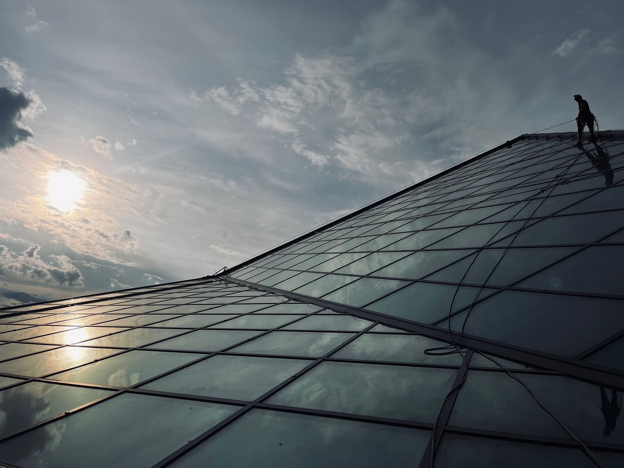 Pracownik na szklanym dachu budynku, zabezpieczony linami. Widok z dołu, słońce odbija się w szybach, pochmurne niebo w tle. Praca na wysokości.
