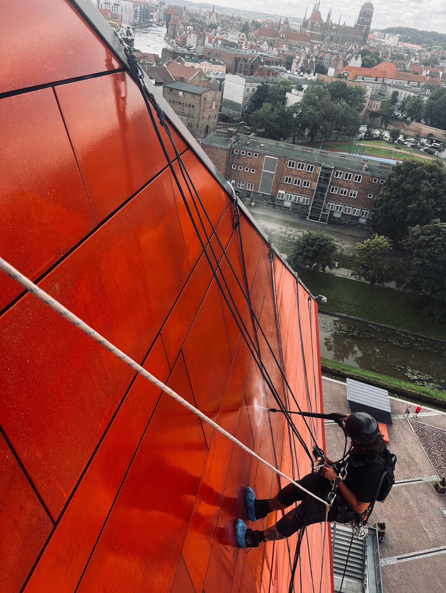 Pracownik w kasku, na linach, czyści czerwoną elewację budynku z użyciem myjki ciśnieniowej. W tle panorama miasta z rzeką i zabytkowymi budowlami.