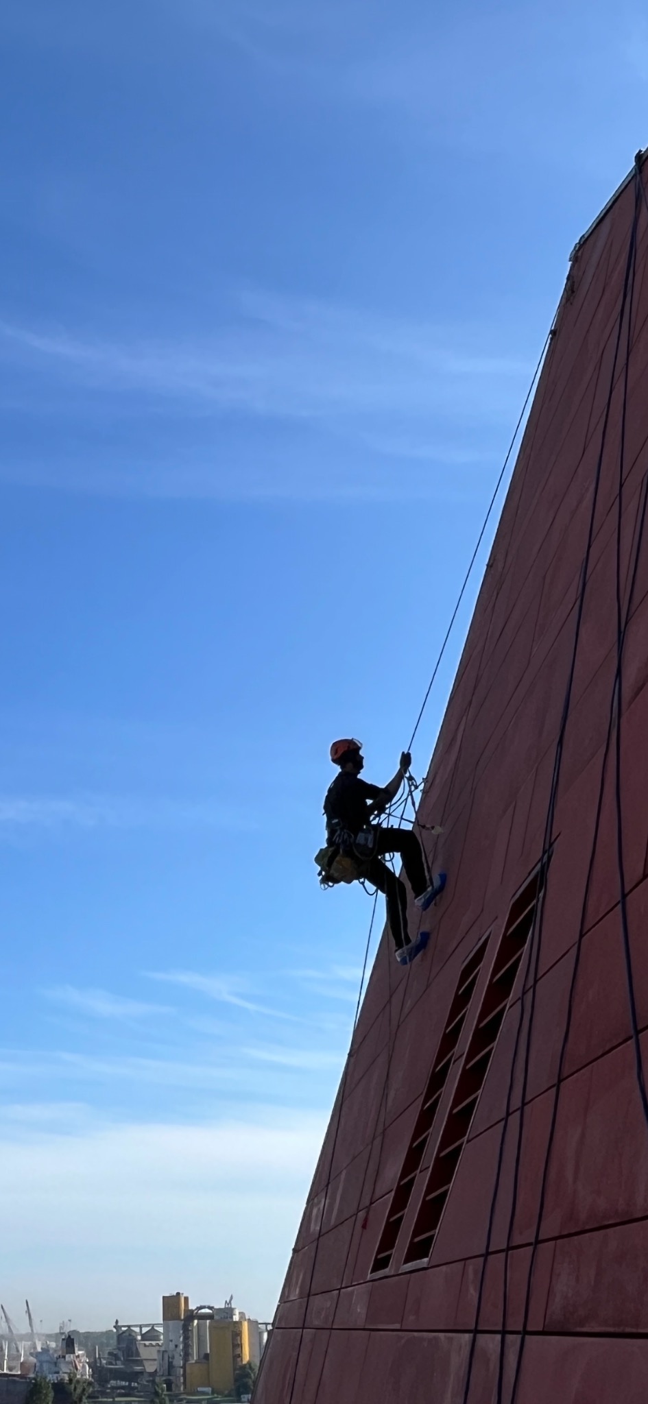 Pracownik w uprzęży, w kasku, na tle błękitnego nieba, zwisa na linie z pionowej ściany budynku o nietypowej architekturze, wykonując prace na wysokości.