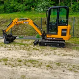 Żółta minikoparka JCB 16c-1 na piaszczystym terenie z zielenią w tle, widoczna z boku, z łyżką skierowaną w lewo, ogrodzenie siatkowe w tle.