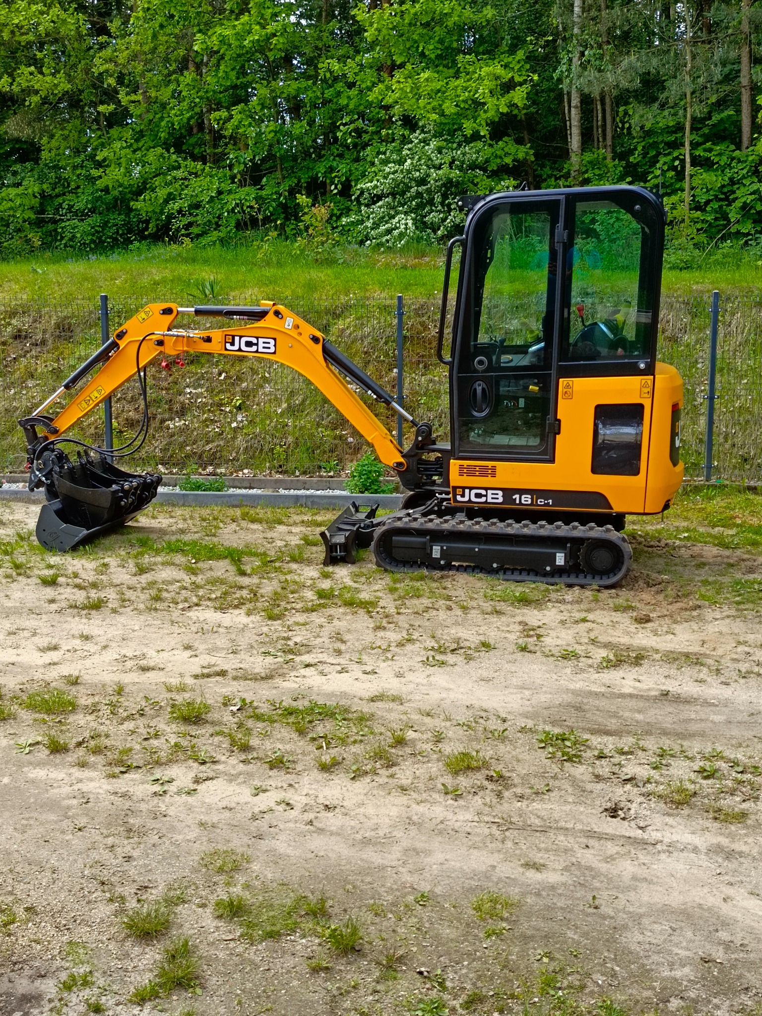 Żółta minikoparka JCB 16c-1 na piaszczystym terenie z zielenią w tle, widoczna z boku, z łyżką skierowaną w lewo, ogrodzenie siatkowe w tle.