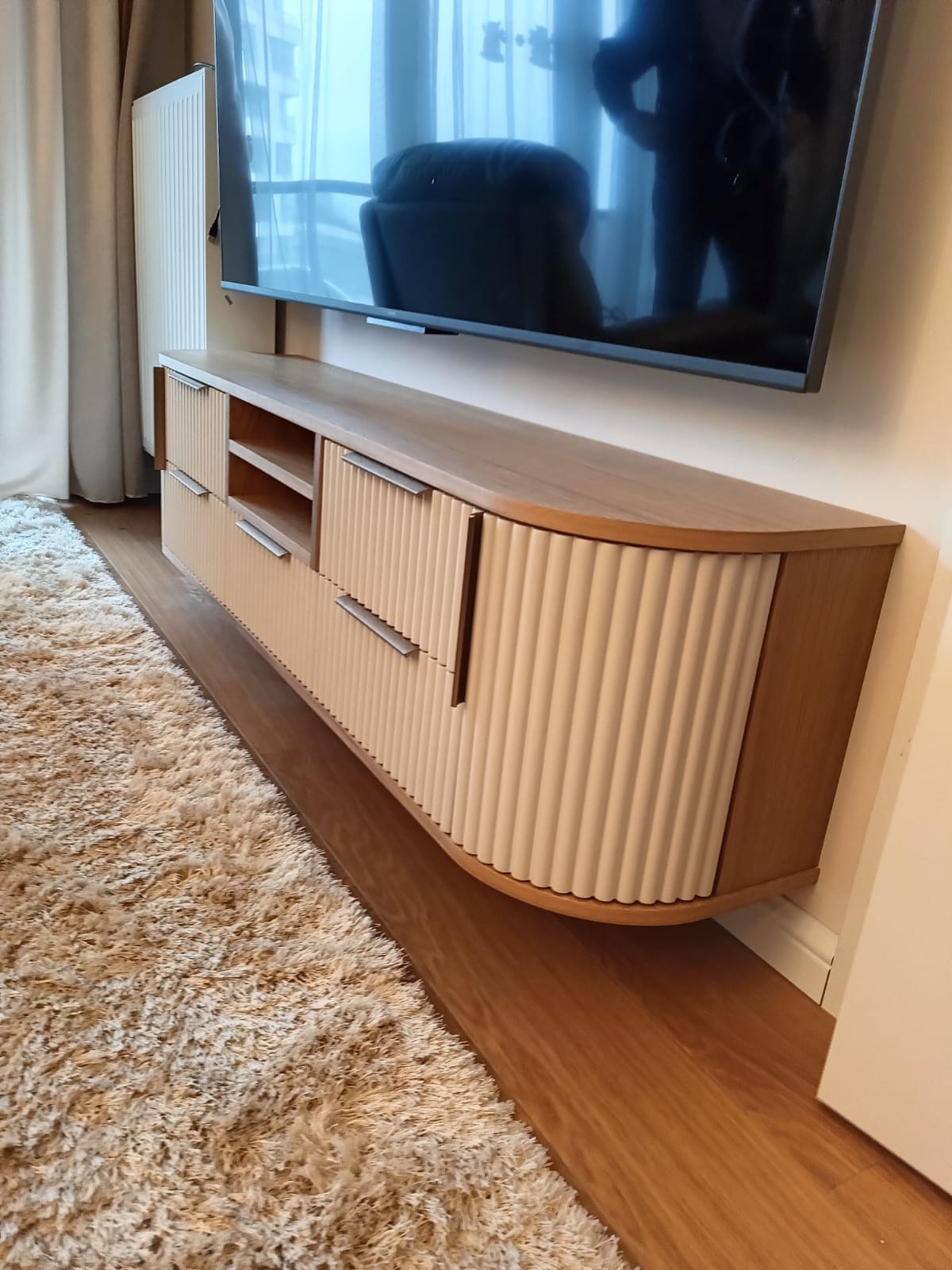 Nowoczesna szafka RTV na wymiar z frezowanymi frontami i drewnianym blatem, zamontowana pod telewizorem w salonie. Widoczny dywan i parkiet.