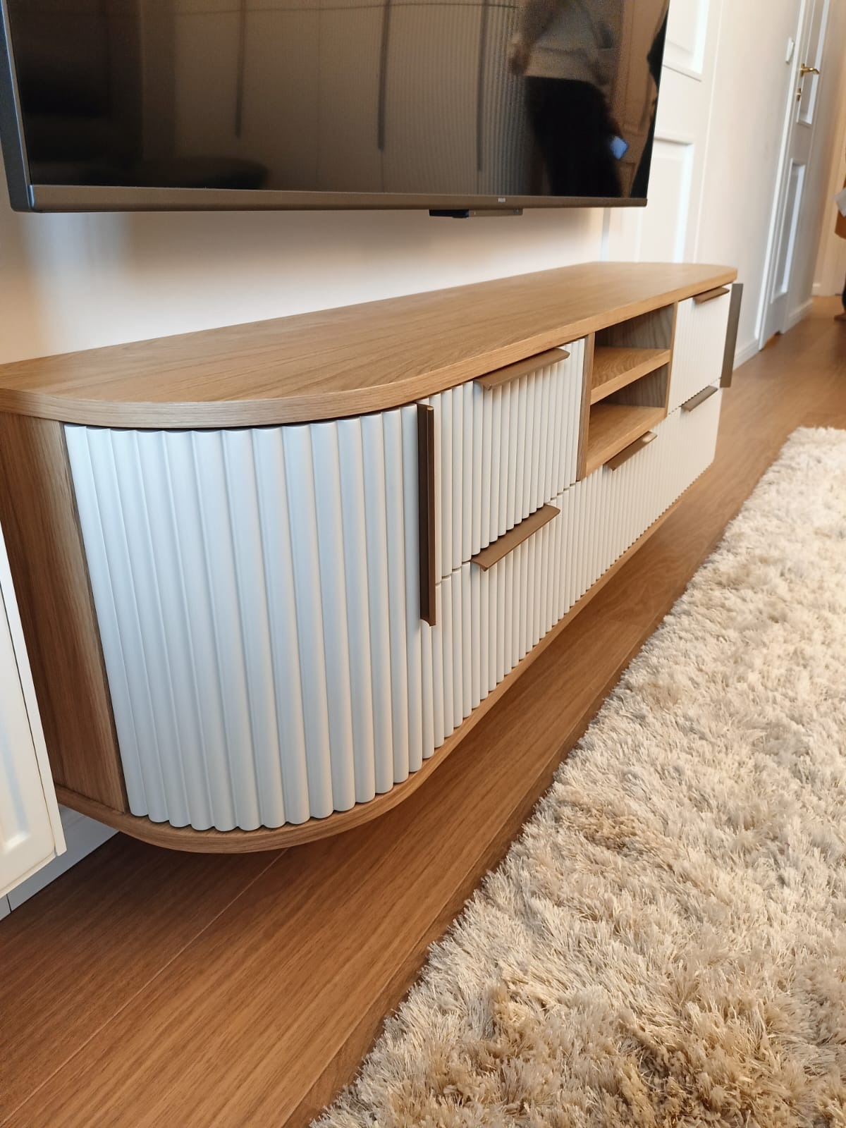 Nowoczesna szafka RTV z frezowanymi frontami w kolorze białym i drewnianym blatem, zawieszona pod telewizorem. Widoczny fragment puszystego dywanu i podłogi.