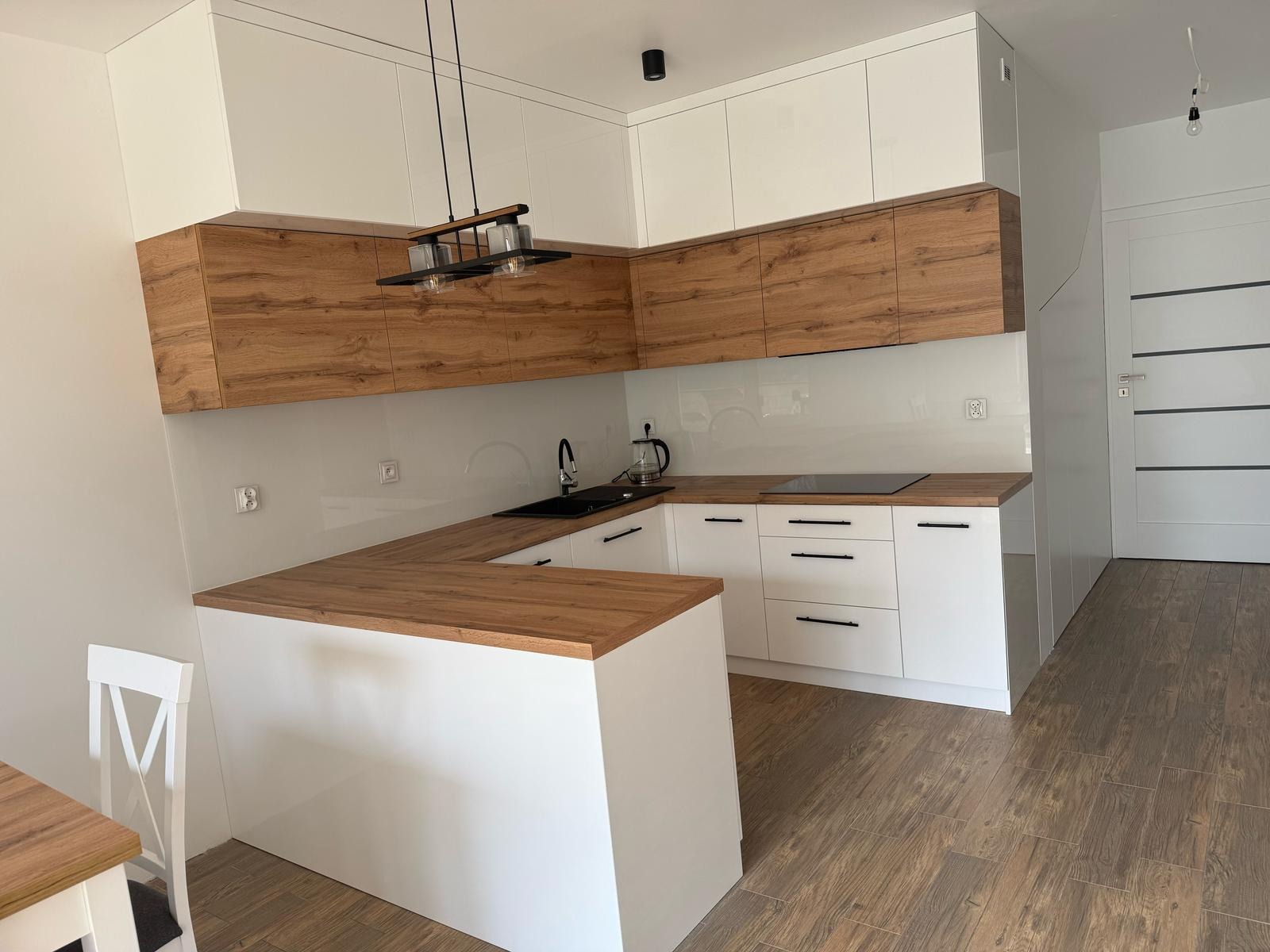 Nowoczesna, narożna kuchnia z białymi frontami i drewnianymi szafkami wiszącymi. Blat z dekorem drewna, czarny zlew i płyta indukcyjna. Oświetlenie punktowe i podłużna lampa.