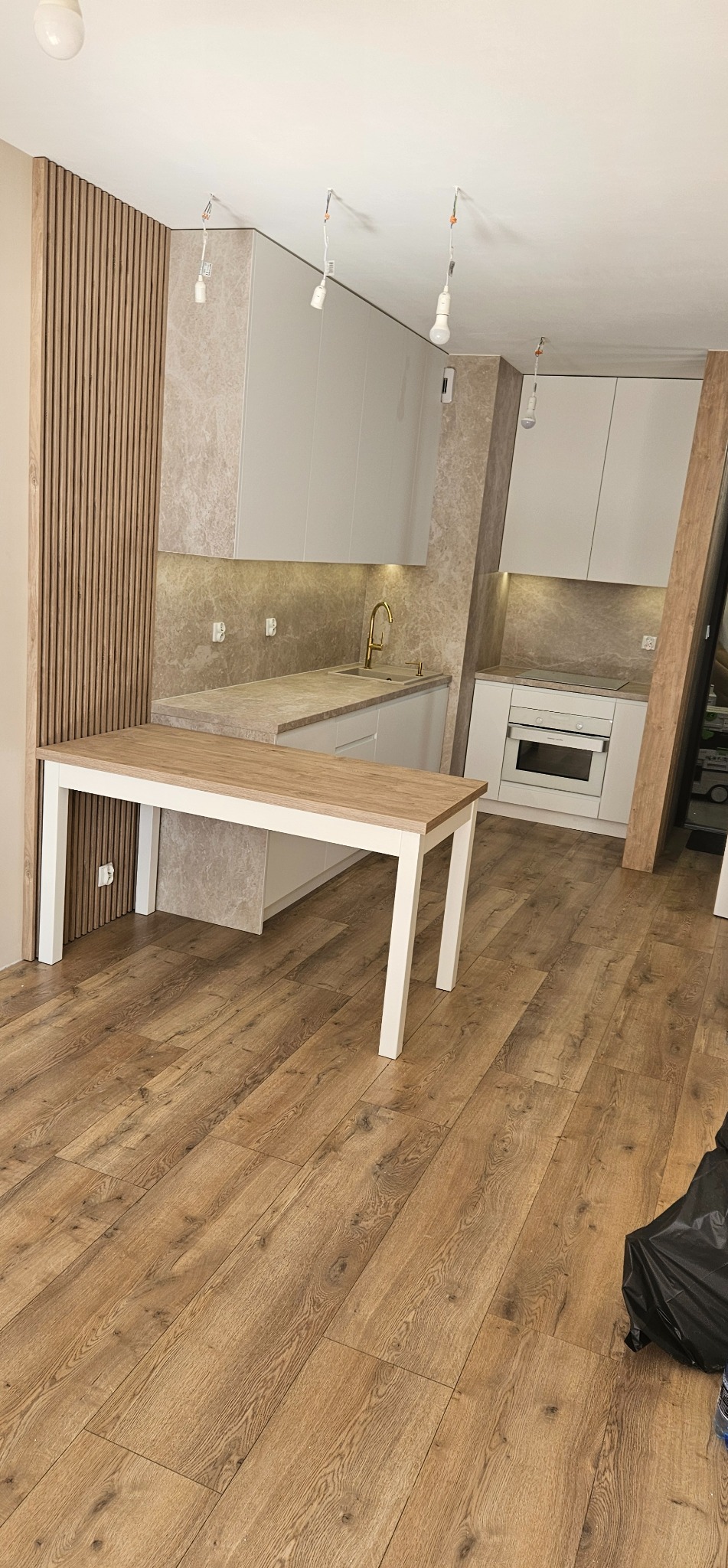 Jasna kuchnia z marmurowym blatem i drewnianym panelem ściennym. Stół z drewnianym blatem i białymi nogami na drewnianej podłodze. Niepodłączone lampy wiszące.