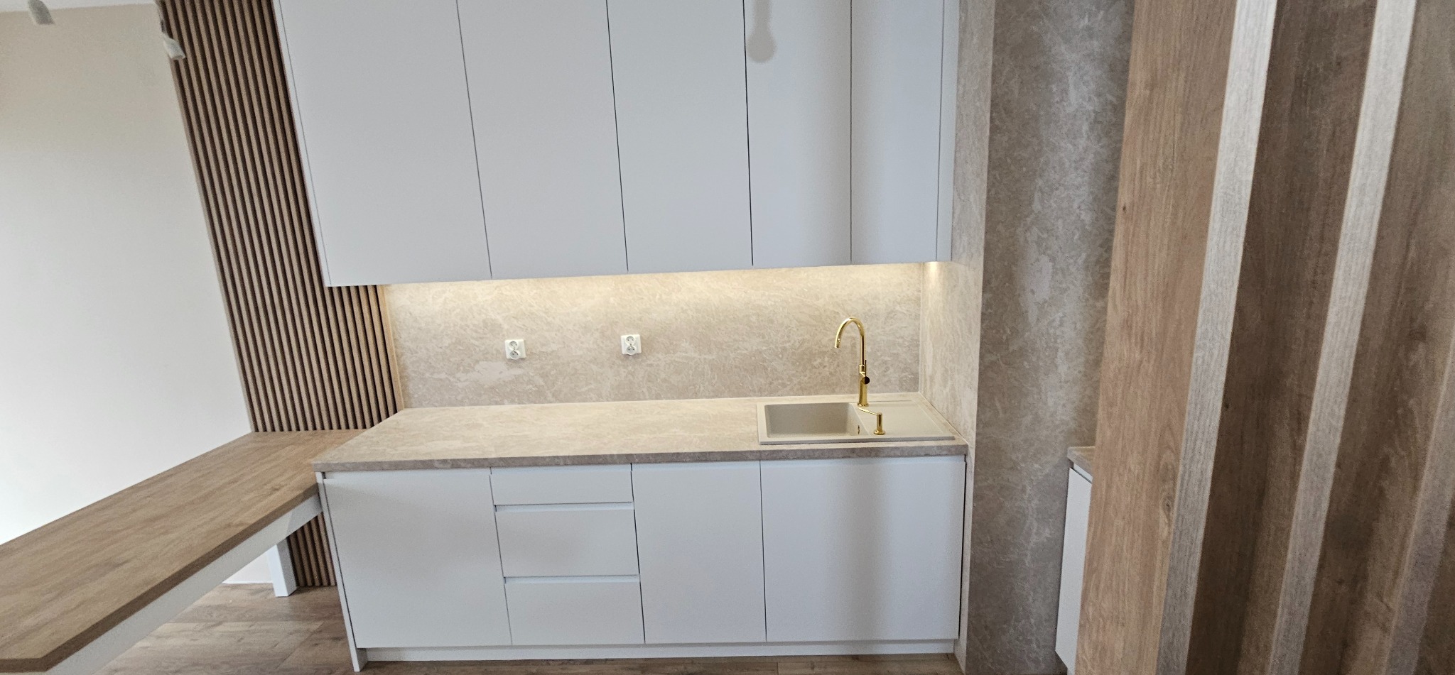 Minimalistyczna kuchnia z białymi szafkami, złotą armaturą i blatem w odcieniach beżu. Na ścianie dekoracyjne panele lamelowe i podświetlenie LED.