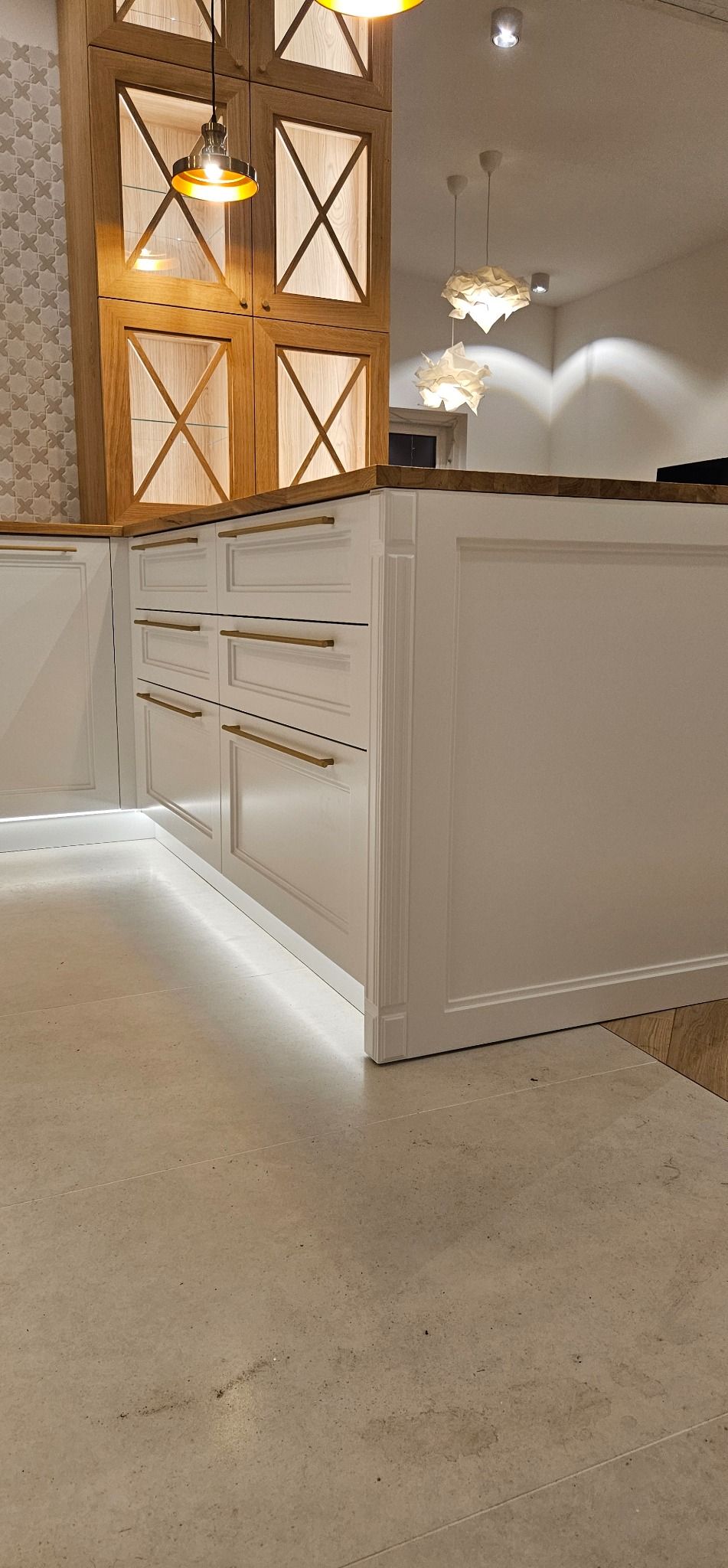 Biała zabudowa kuchenna z szufladami i złotymi uchwytami, podświetleniem LED u dołu i drewnianymi szafkami wiszącymi w tle. Element wyspy kuchennej.