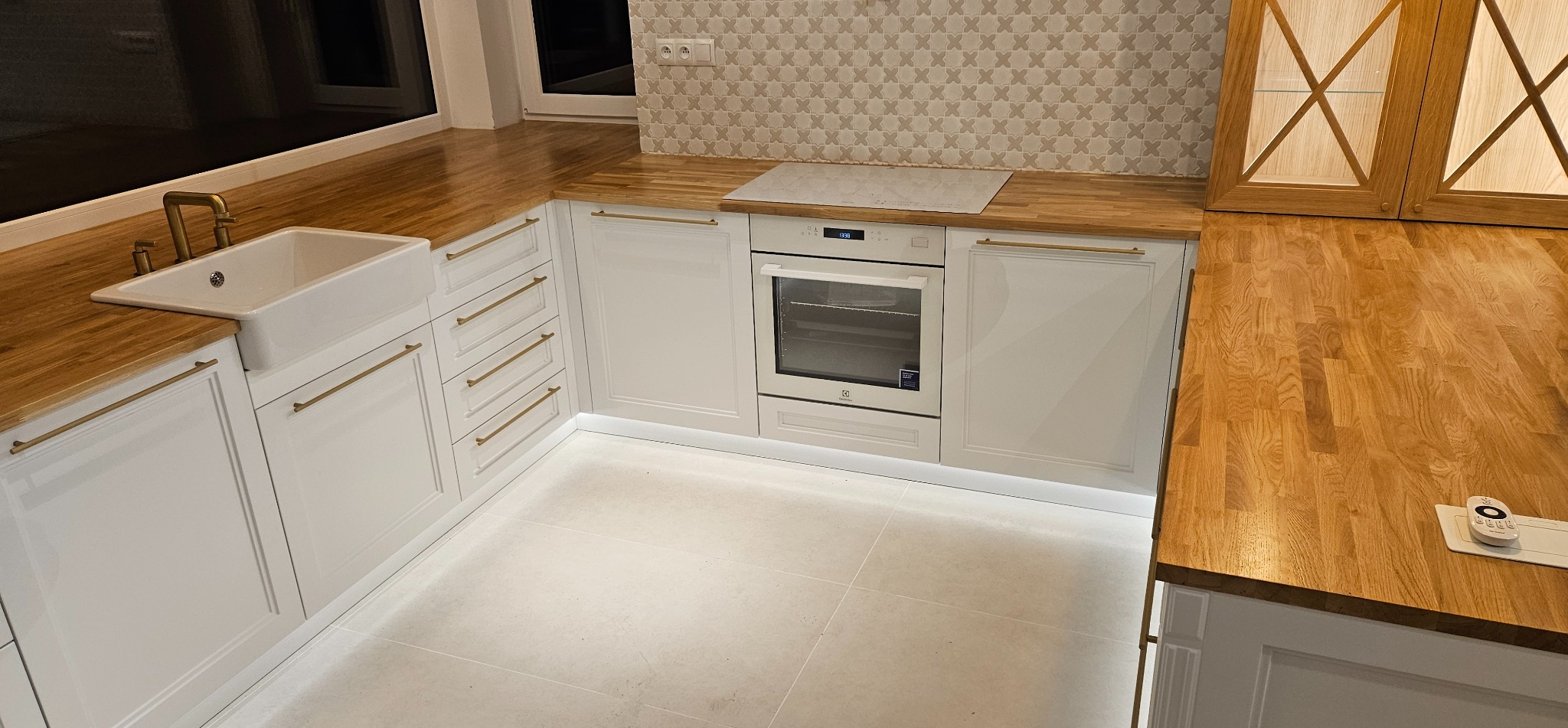 Jasna kuchnia z drewnianym blatem: białe szafki z frezowanymi frontami, złote uchwyty, ceramiczny zlew i subtelne podświetlenie LED.