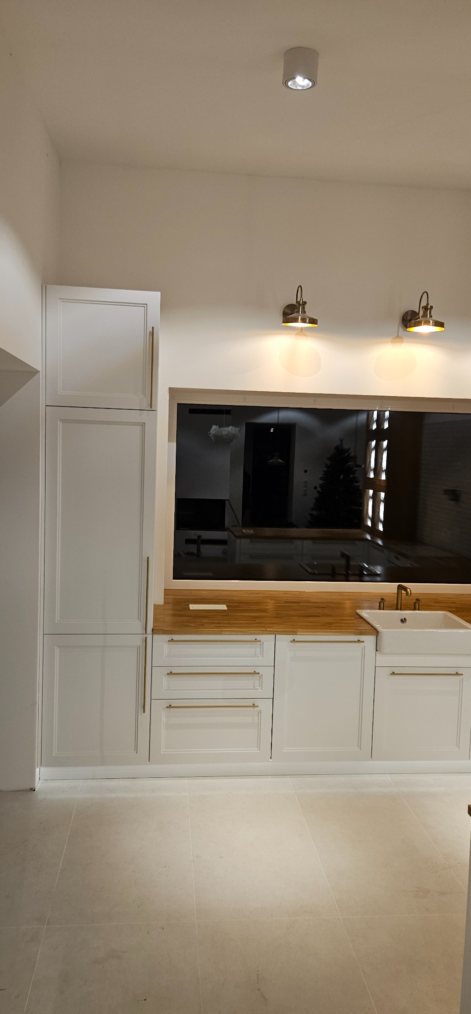 Biała zabudowa kuchenna z drewnianym blatem i złotymi uchwytami, oświetlona lampami ściennymi. W tle widoczne odbicie wnętrza z choinką.