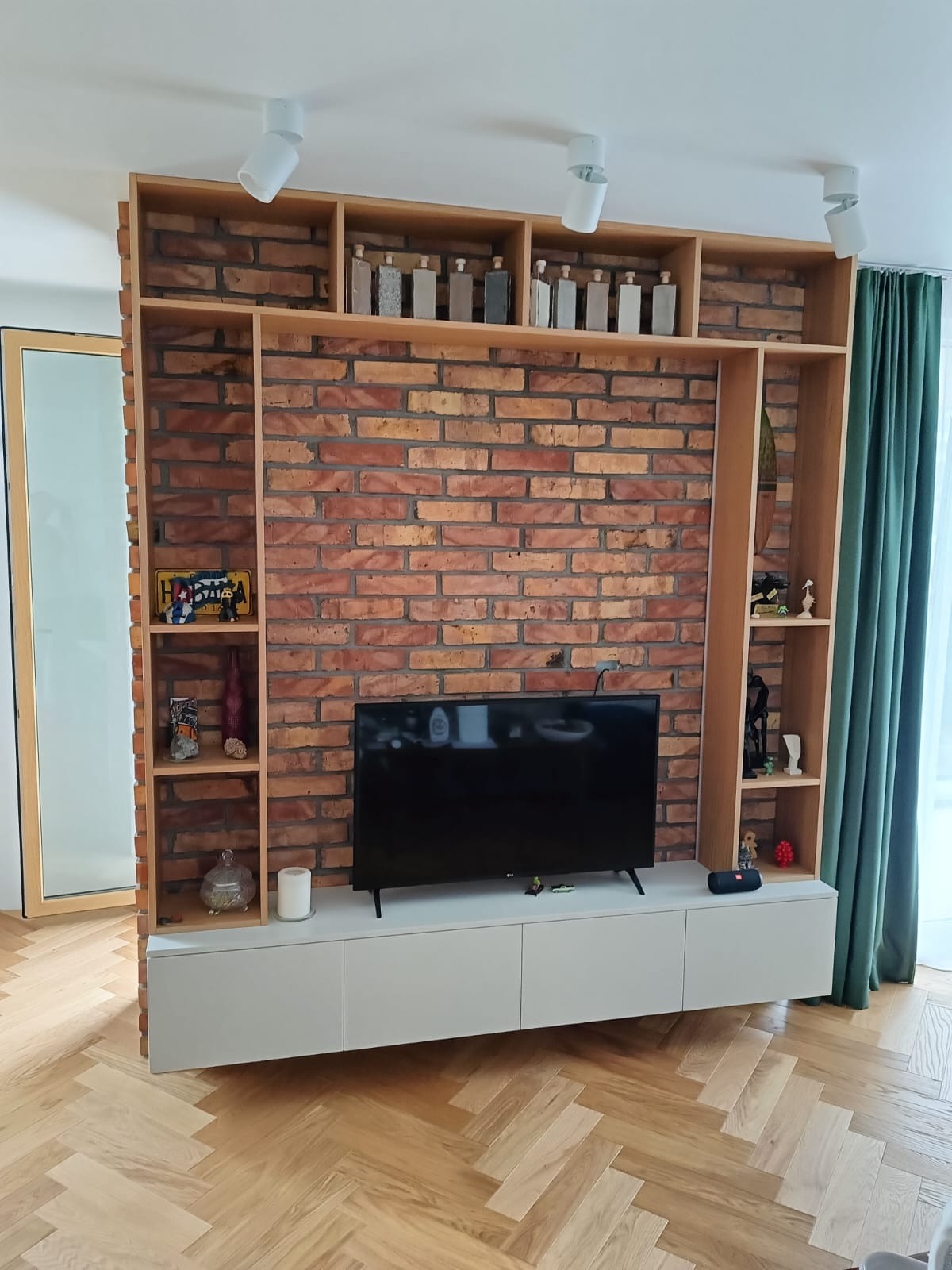 Szafka RTV z ceglaną ścianą i drewnianymi półkami w salonie. Dekoracyjne butelki na górnych półkach, parkiet w jodełkę. Nowoczesny design.