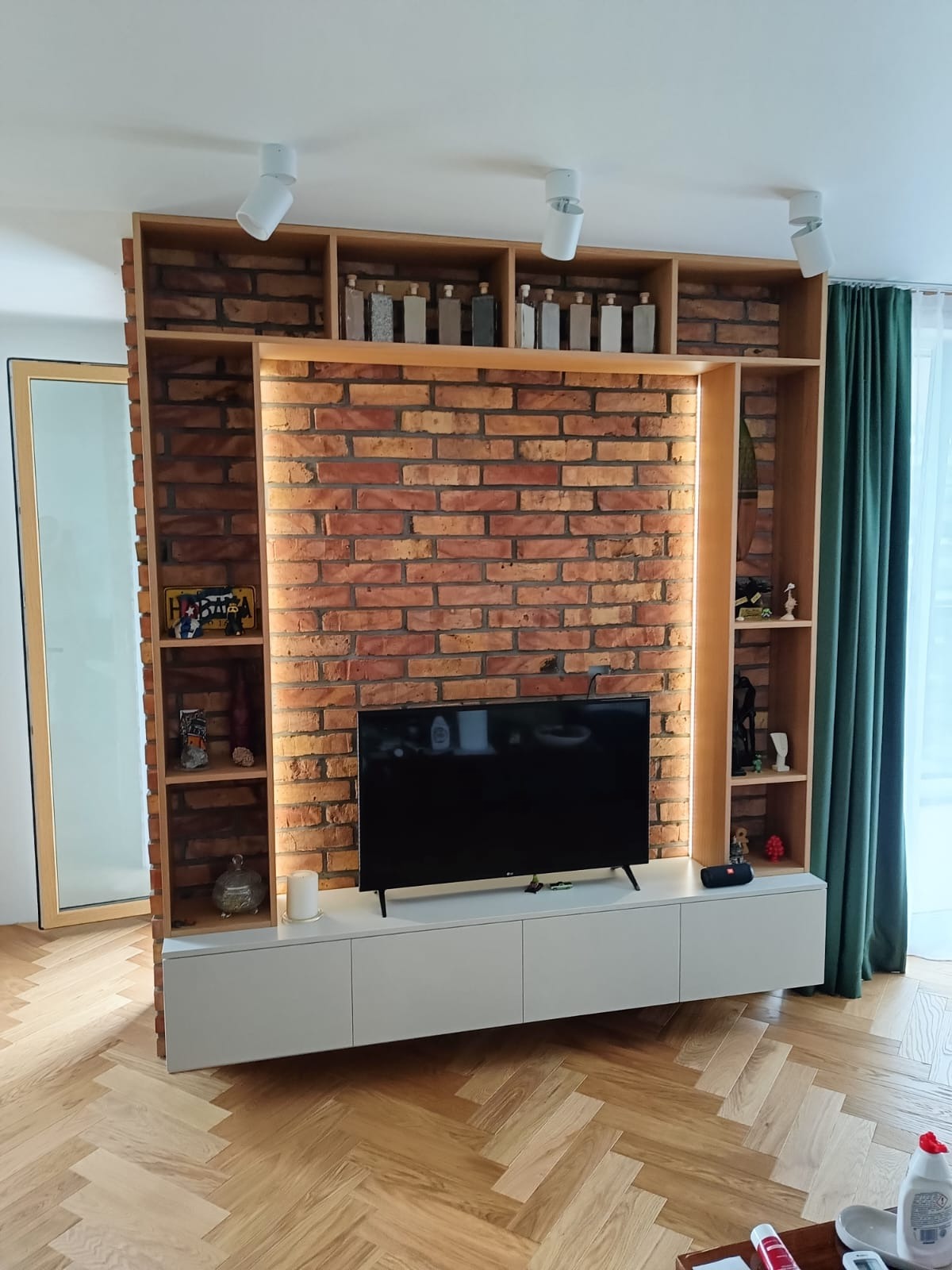 Regał z ceglaną ścianą i podświetleniem LED, z półkami na dekoracje i szafką RTV w salonie z parkietem w jodełkę. Górne półki z pojemnikami.