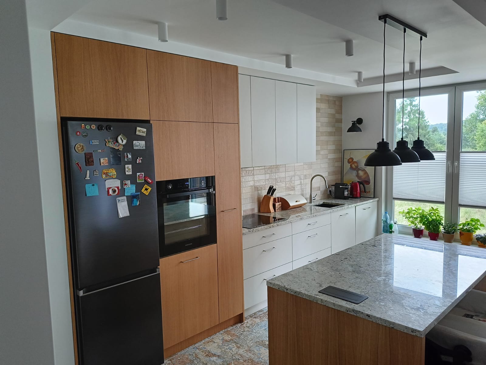 Nowoczesna kuchnia z drewnianymi i białymi frontami, czarną lodówką z magnesami i wyspą z blatem w plamki, oświetlona wiszącymi lampami.