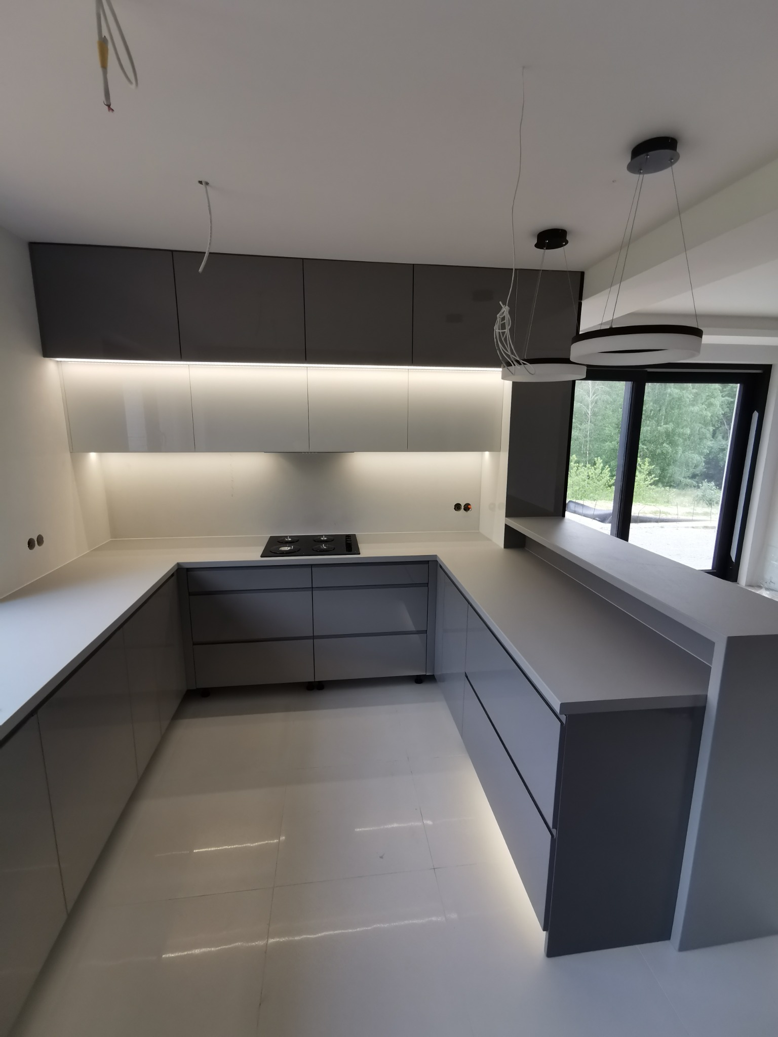 Nowoczesna kuchnia w odcieniach szarości z białymi elementami, oświetleniem LED podszafkowym i wyspą z widokiem na zieleń za oknem. Minimalistyczny design i geometryczne lampy.