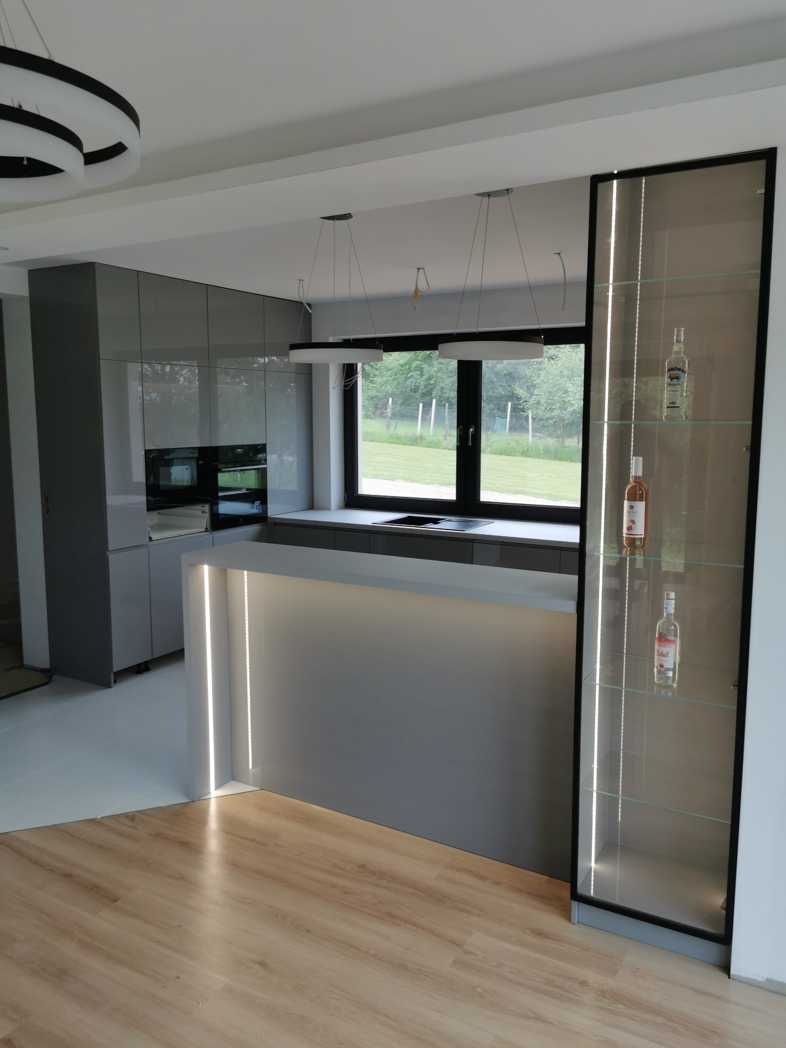Nowoczesna kuchnia z szarymi frontami, oświetloną wyspą i przeszkloną witryną z czarną ramą. Minimalistyczne lampy wiszące. Widok na zewnątrz przez okno.