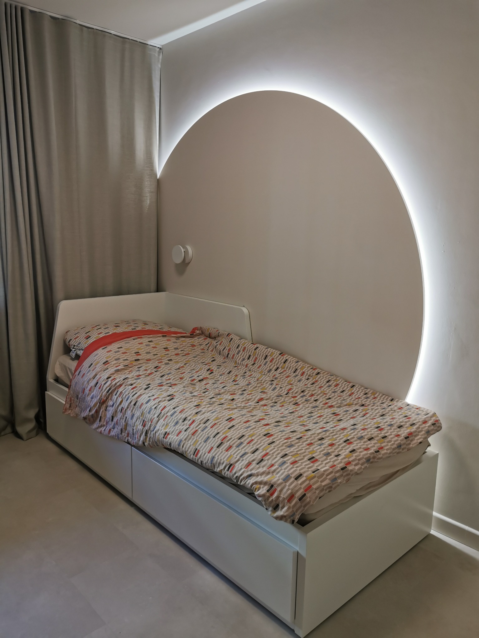 Białe łóżko z szufladami i okrągłym zagłówkiem z podświetleniem LED, wzorzysta pościel, szare zasłony w tle. Nowoczesny design sypialni dziecięcej.