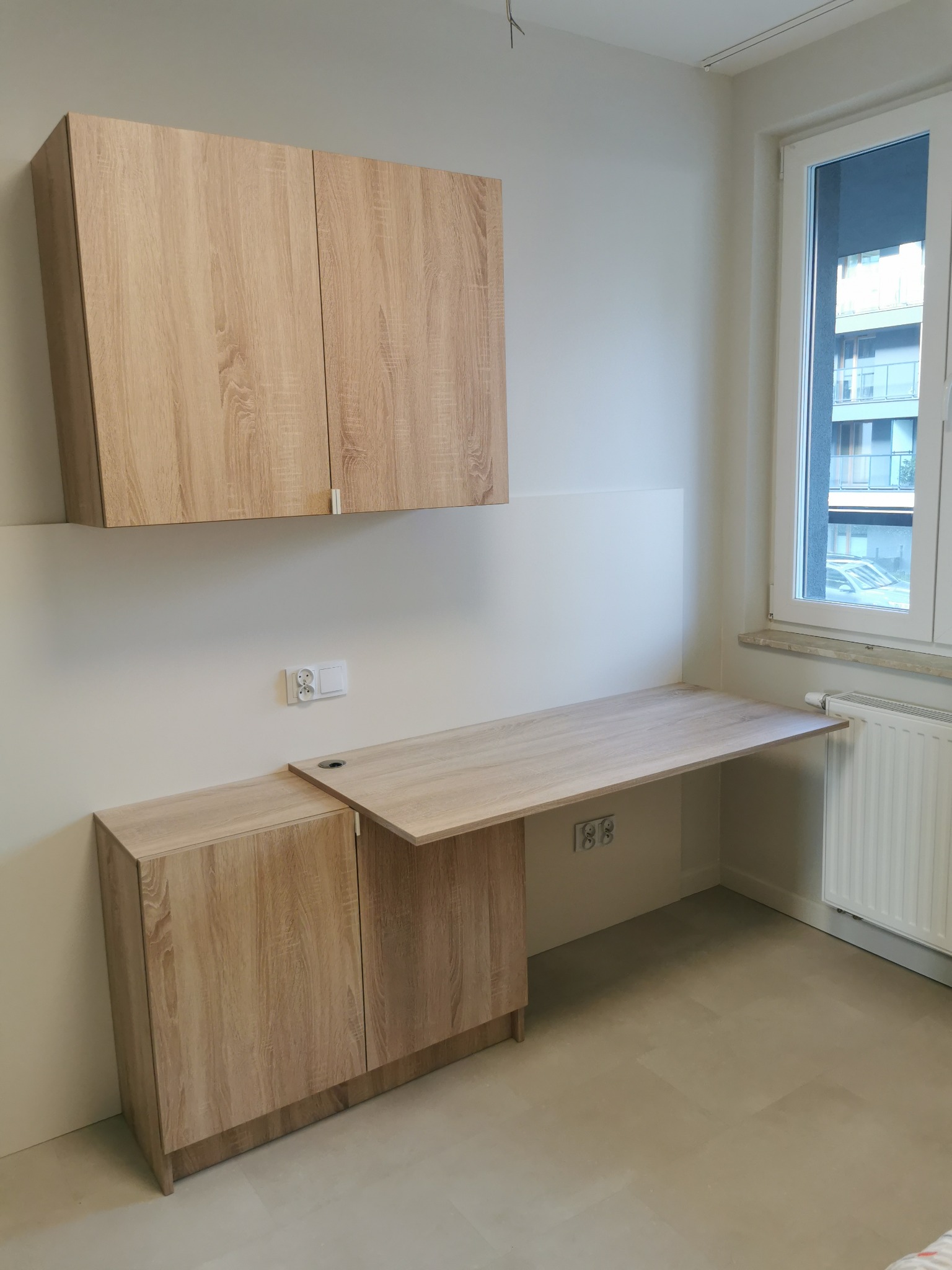 Jasne drewniane szafki wiszące nad biurkiem z blatem, widoczne gniazdka elektryczne i okno z widokiem na budynek wielorodzinny.