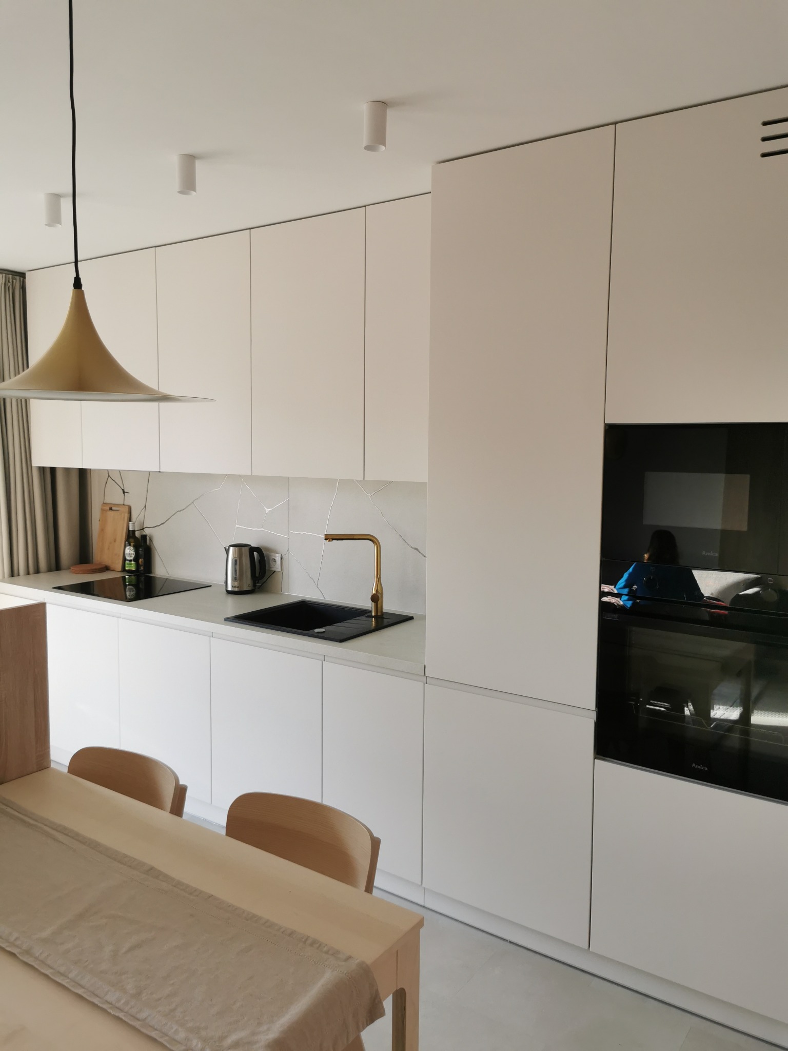 Minimalistyczna, biała kuchnia z blatem imitującym marmur, złotą armaturą i czarnym zlewem. Widoczny stół z krzesłami i lampa w kształcie stożka.