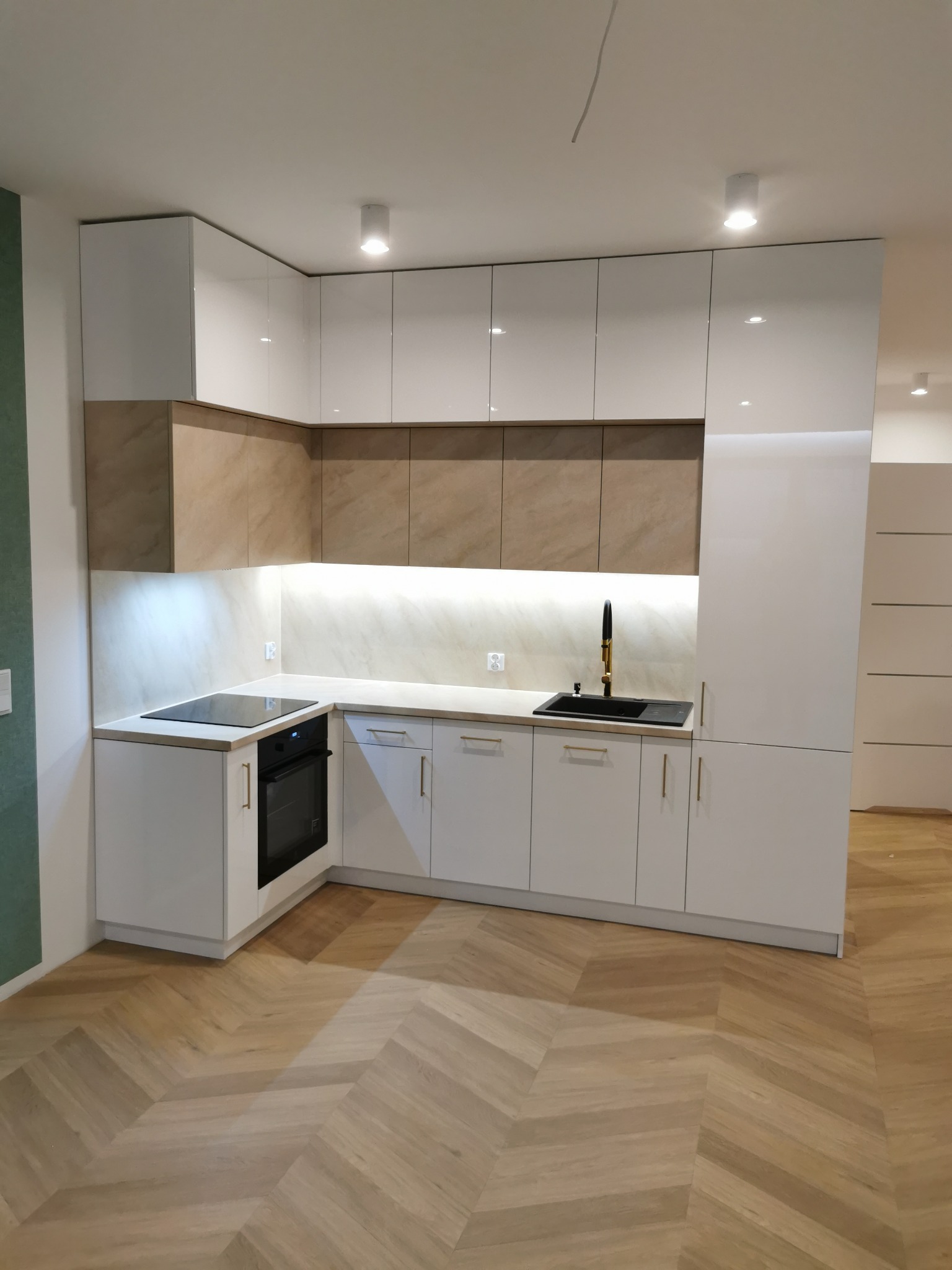 Nowoczesna kuchnia na wymiar w Kalwarii Zebrzydowskiej. Białe fronty szafek dolnych i górnych, beżowe panele nad blatem, złota armatura, parkiet w jodełkę.