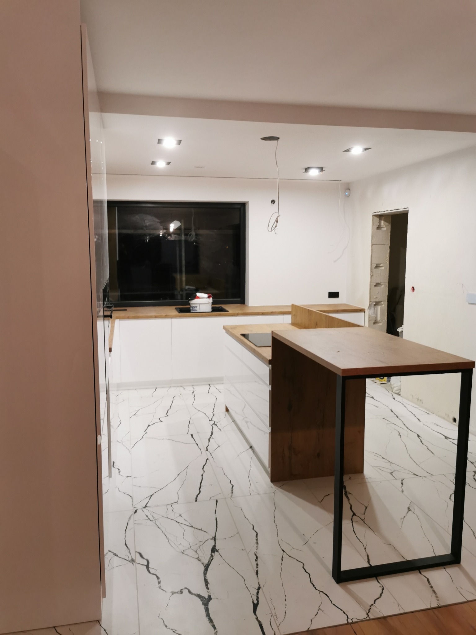 Nowoczesna kuchnia z wyspą na tle białych ścian i marmurowej podłogi. Widoczne minimalistyczne szafki i czarne, duże okno. Częściowo widoczne niedokończone wykończenia.