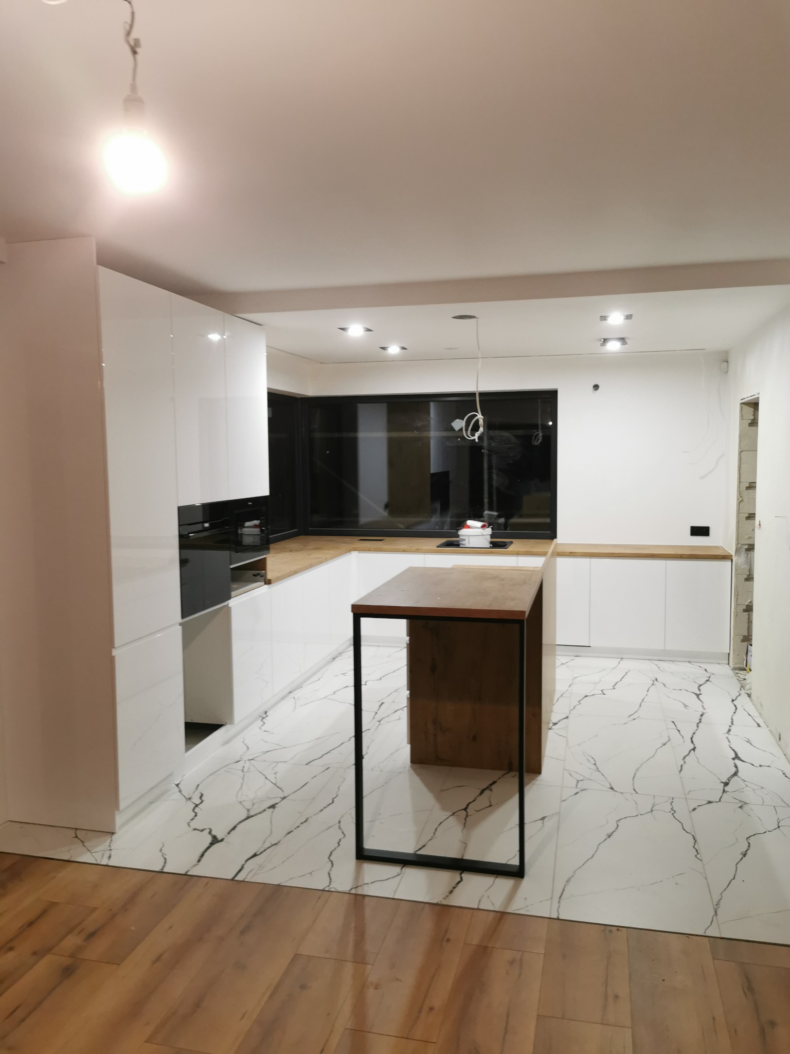 Nowoczesna kuchnia z białymi frontami, drewnianym blatem i wyspą z czarnymi nogami. Marmurowe płytki podłogowe i drewniana podłoga w sąsiednim pomieszczeniu.