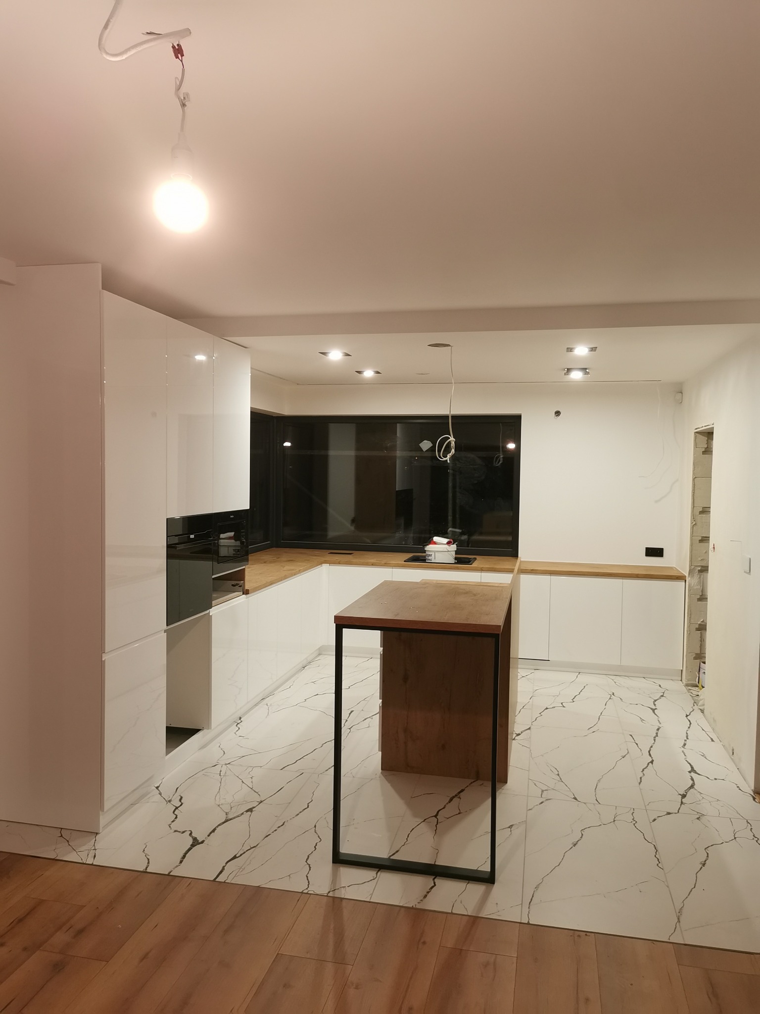 Nowoczesna, biała kuchnia z drewnianym blatem i wyspą na marmurowej podłodze. Widoczne niedokończone prace instalacyjne, surowe wykończenie ścian.